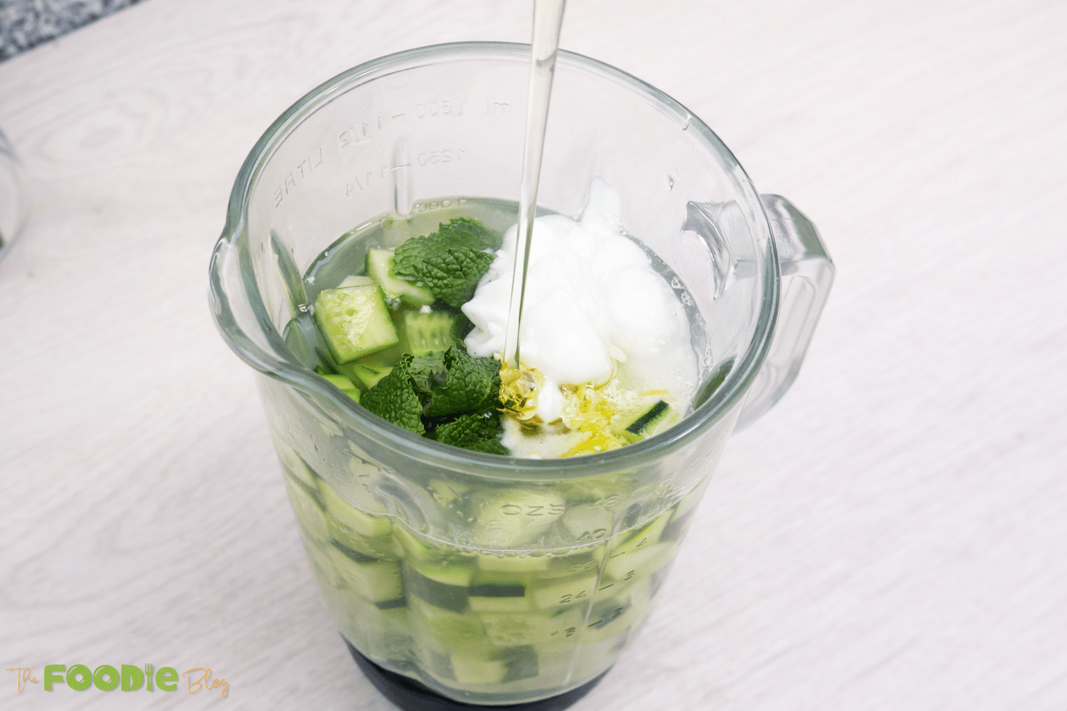 Honey being poured into a blender jar with cucumber, mint, yogurt, and lemon zest