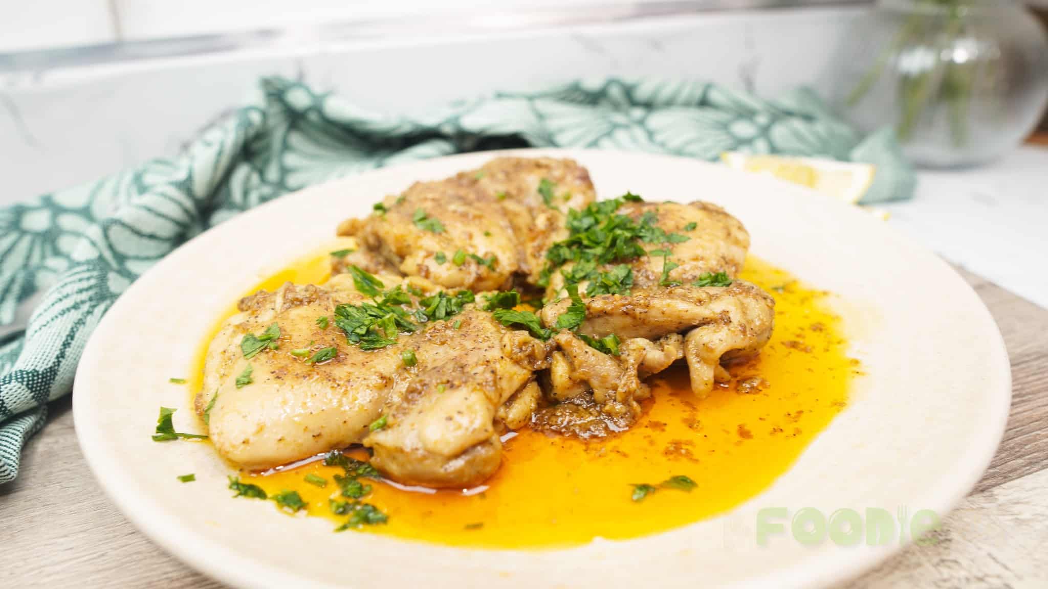 Garlic chicken thighs on a white plate with golden sauce, herbs, and a lemon wedge in the background