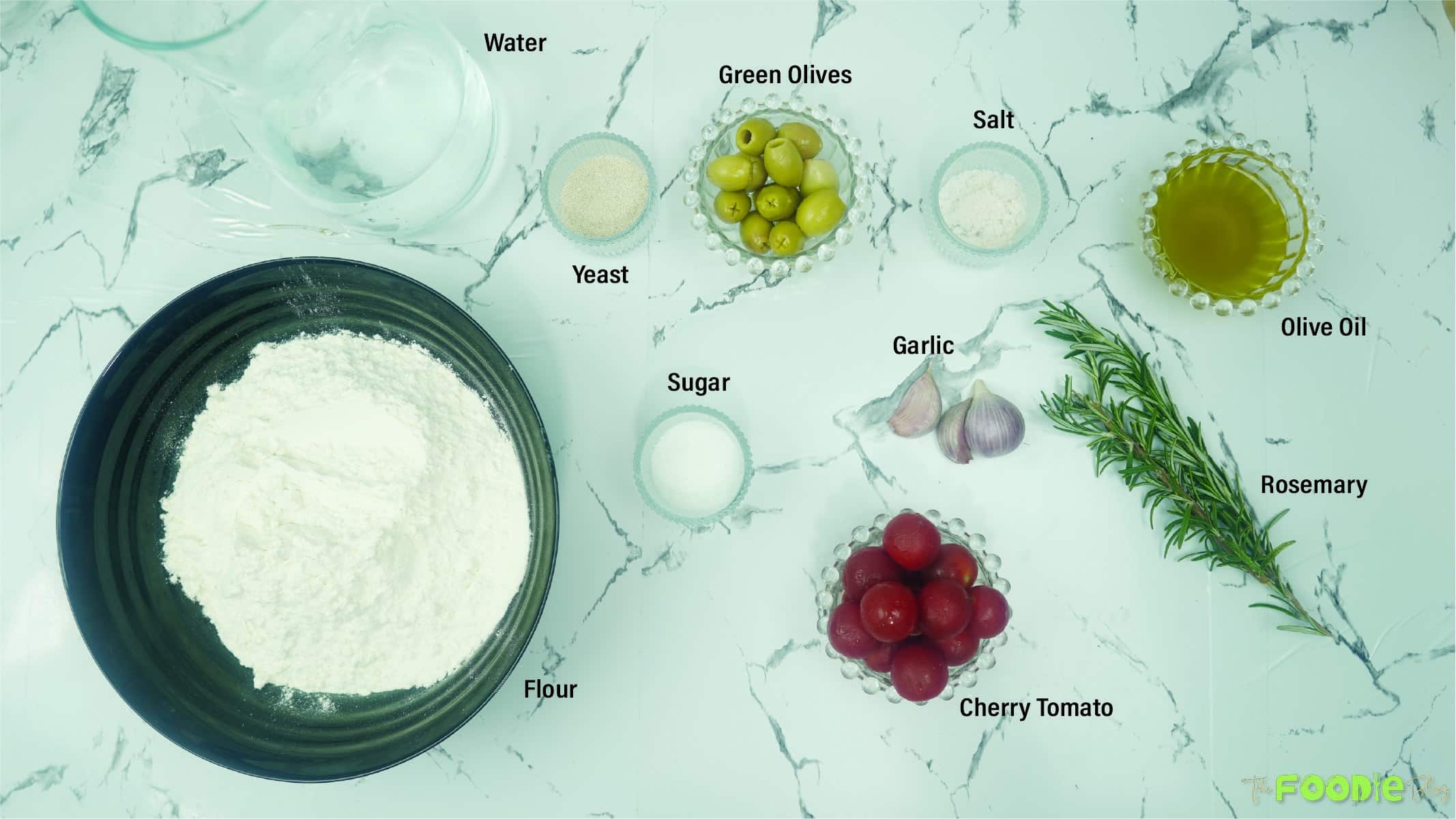 Overhead view of flour, water, yeast, sugar, salt, olive oil, olives, cherry tomatoes, garlic, and rosemary