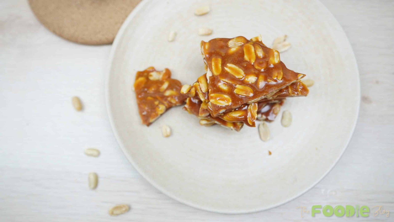 Peanut brittle pieces stacked on a plate with a few peanuts scattered around Caption: Finished peanut brittle ready to serve