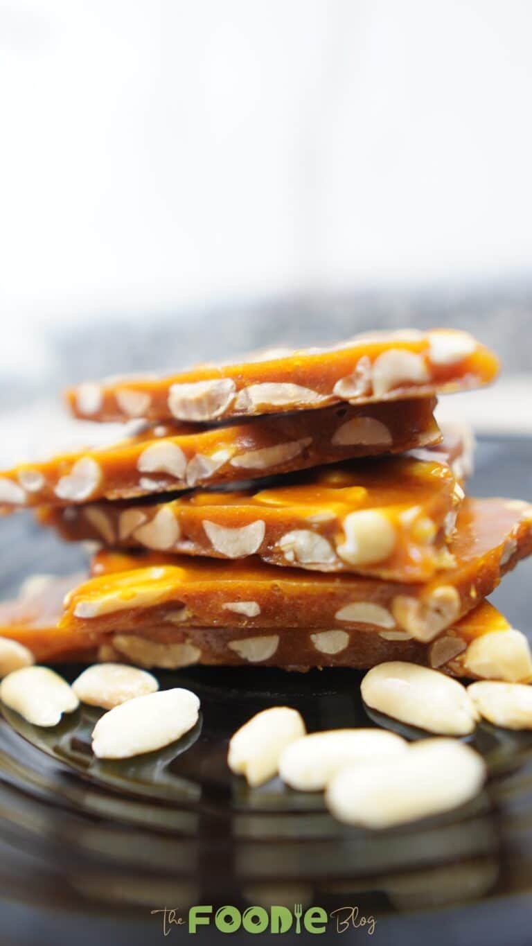 Stack of glossy peanut brittle pieces on a dark plate with peanuts around it Caption: Crisp, buttery peanut brittle with roasted peanuts