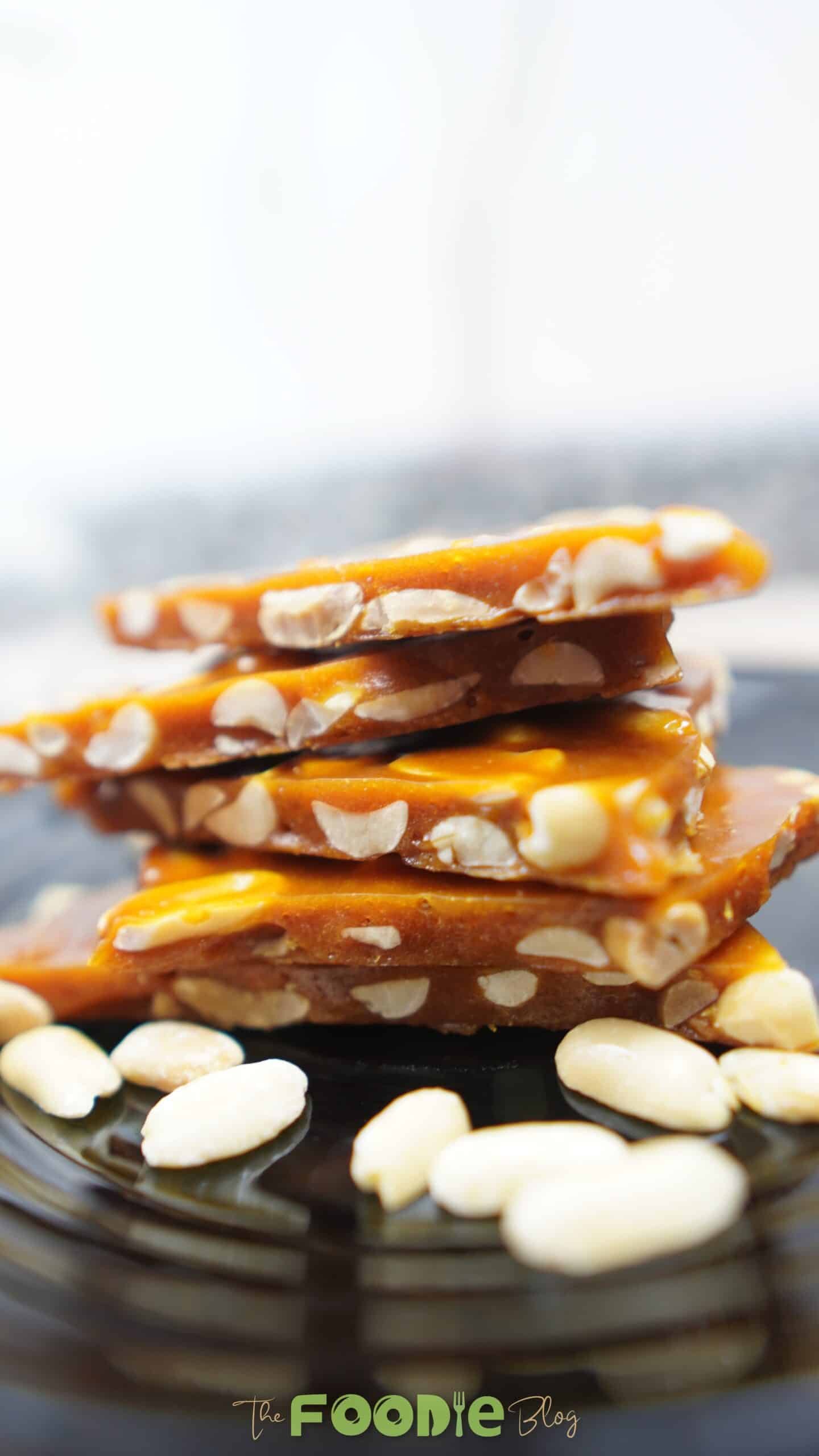 Stack of glossy peanut brittle pieces on a dark plate with peanuts around it Caption: Crisp, buttery peanut brittle with roasted peanuts