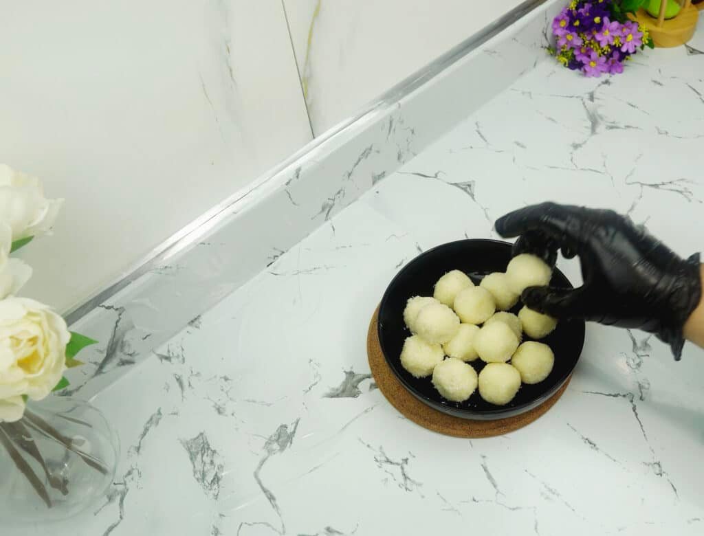 Hand placing coconut-coated balls into a black bowl on the counter