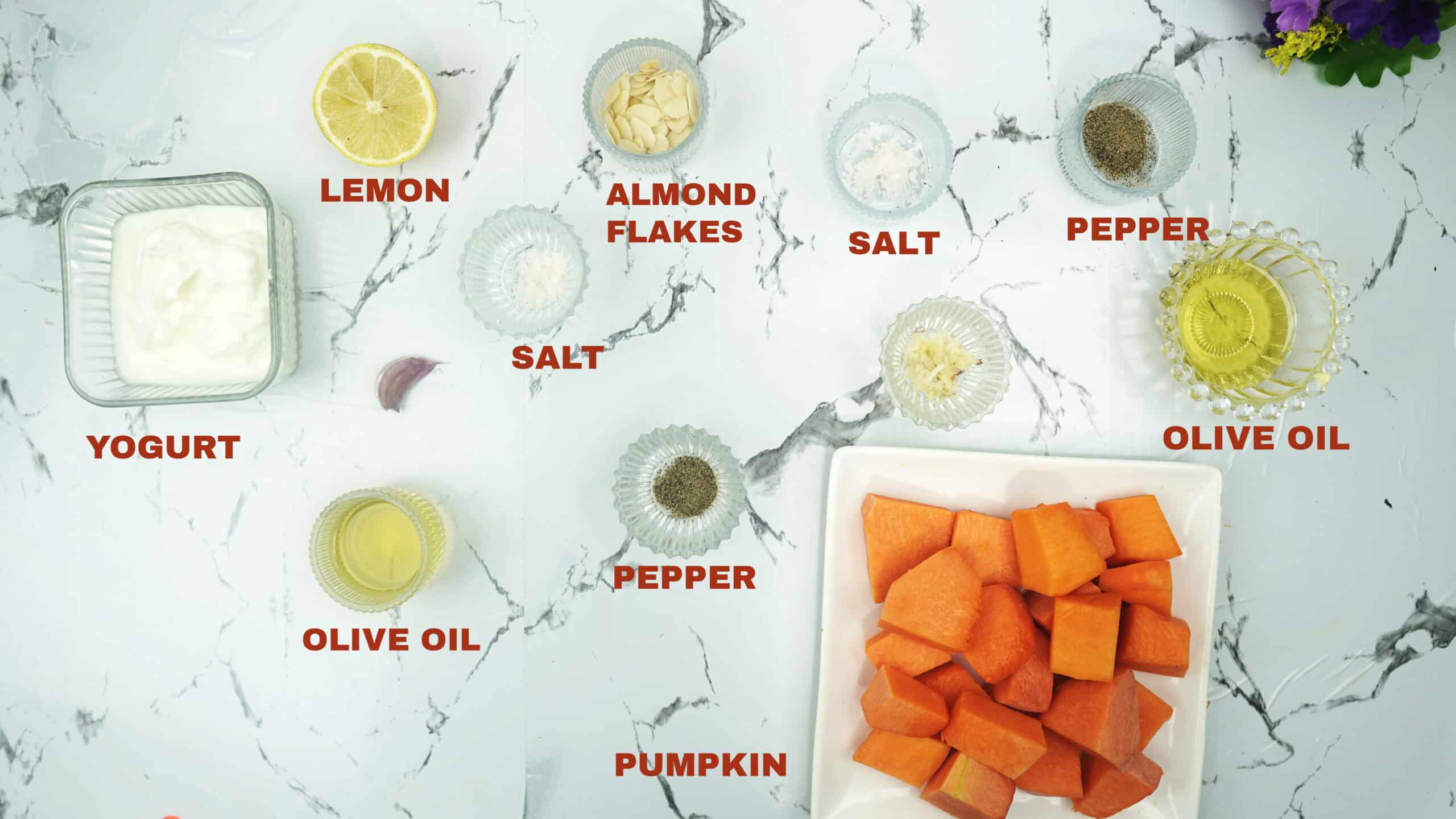 Labeled ingredients on a white counter including pumpkin, yogurt, lemon, olive oil, garlic, salt, pepper, and almond flakes