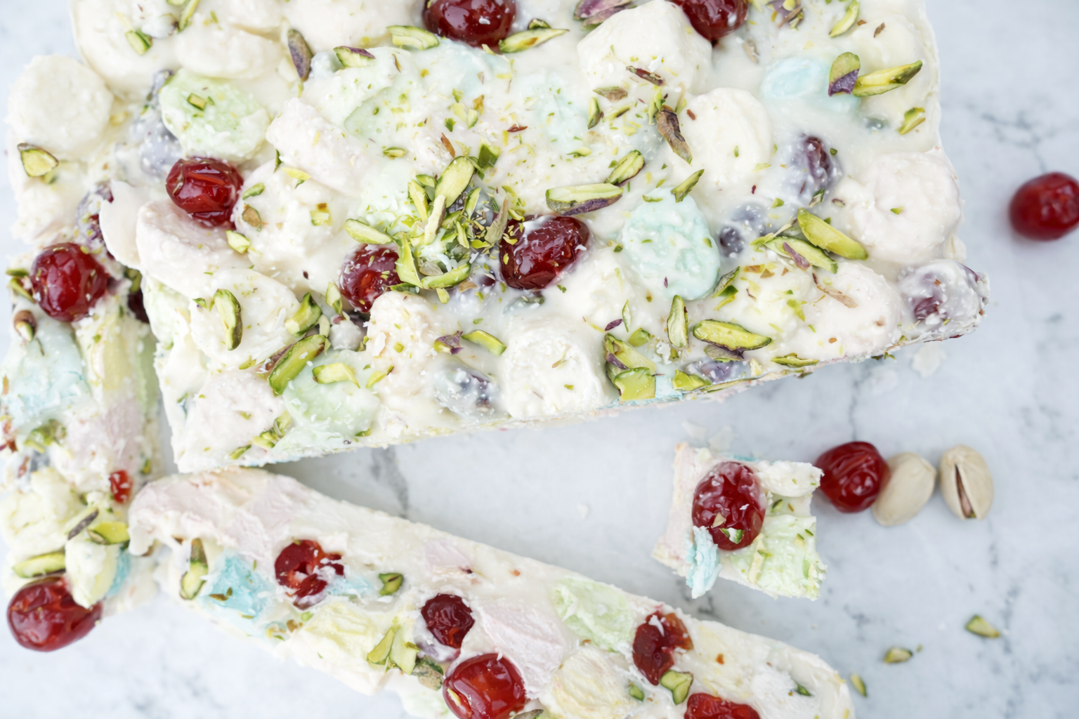 White Christmas rocky road slab topped with glacé cherries and pistachios on a marble background