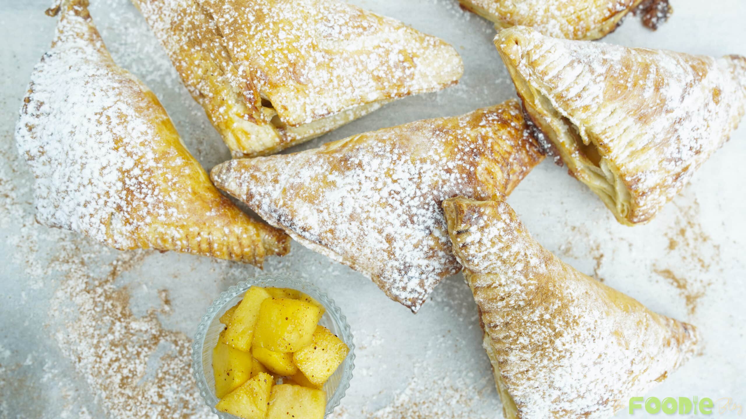 Finished apple turnovers dusted with powdered sugar on a tray with a small bowl of apple filling