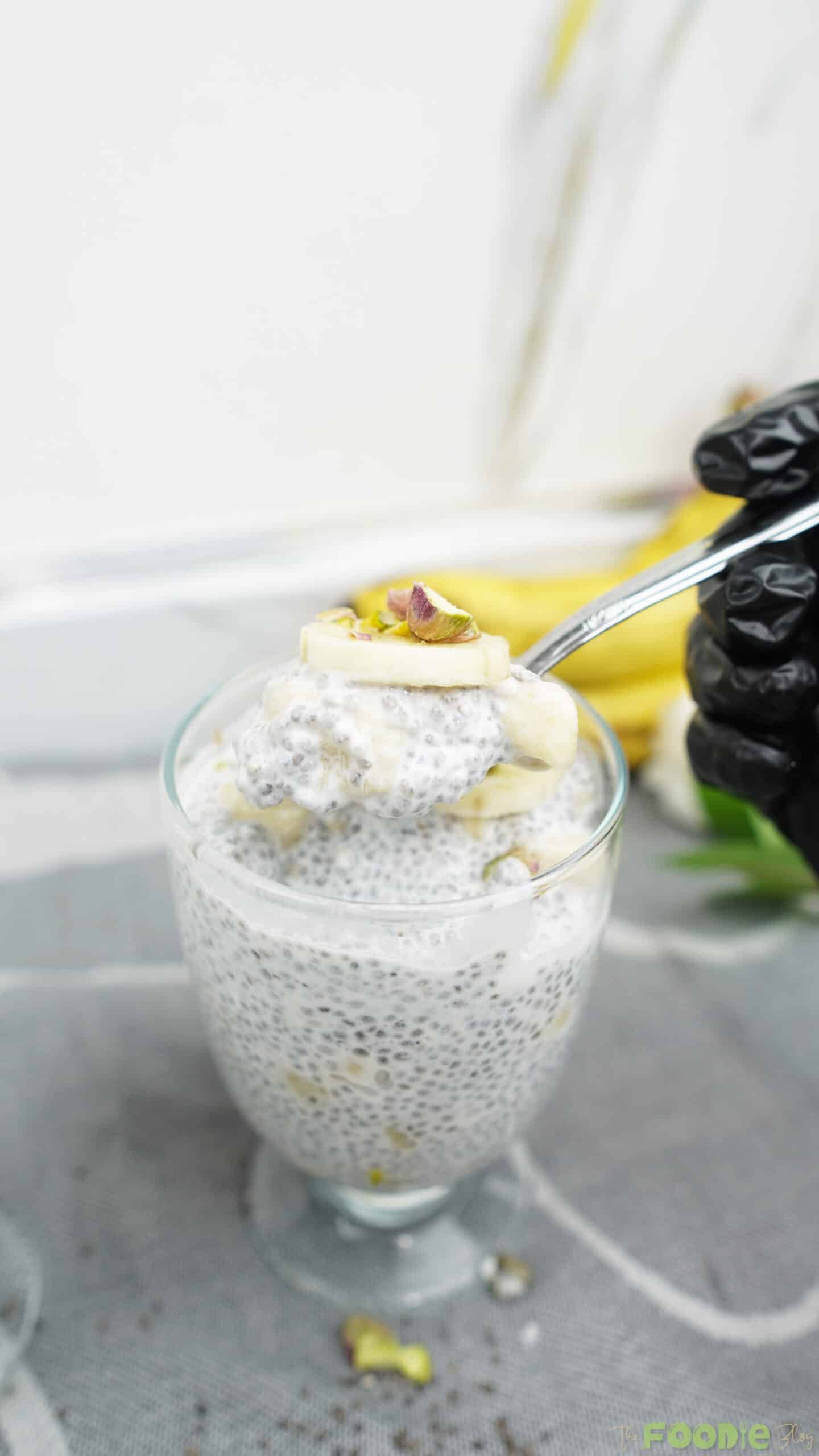 A spoon lifting a bite of chia pudding with banana from a glass, with bananas in the background.