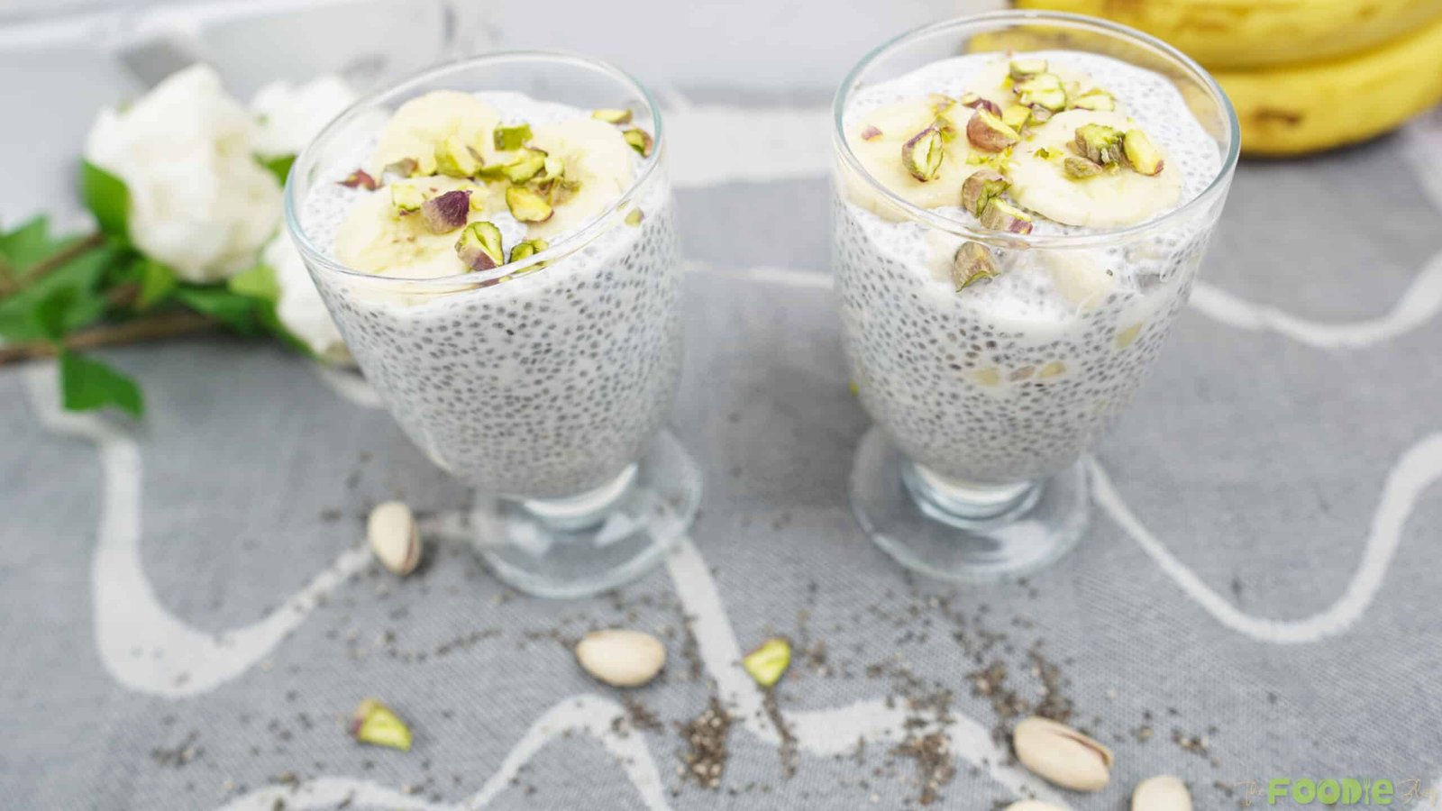 Two glasses of chia pudding with banana slices and pistachios on top, styled on a table.