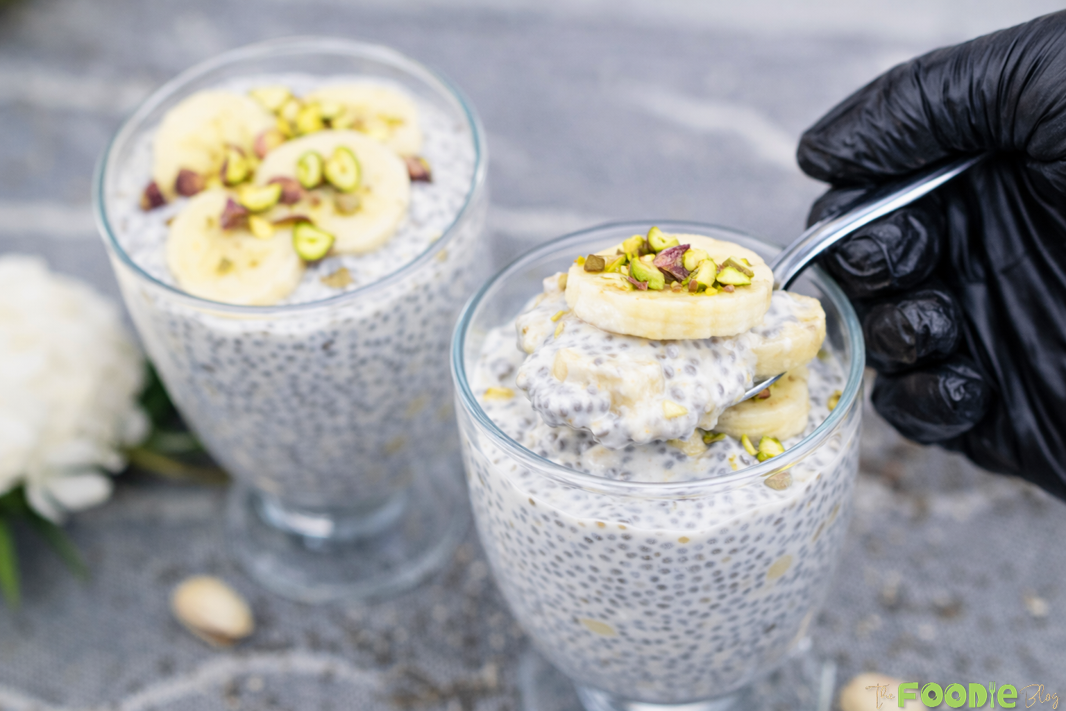 Two glasses of banana chia seed pudding with banana slices and pistachios, with a spoon scooping.