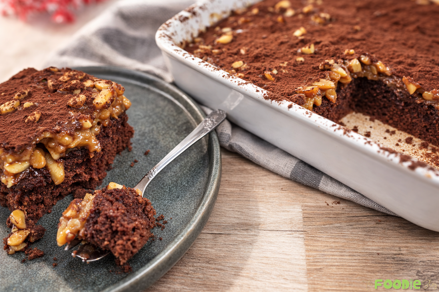 A slice of Caramel Peanut Delight Cake served on a plate with a spoon, with the remaining cake in a baking dish topped with caramel peanuts and cocoa powder.