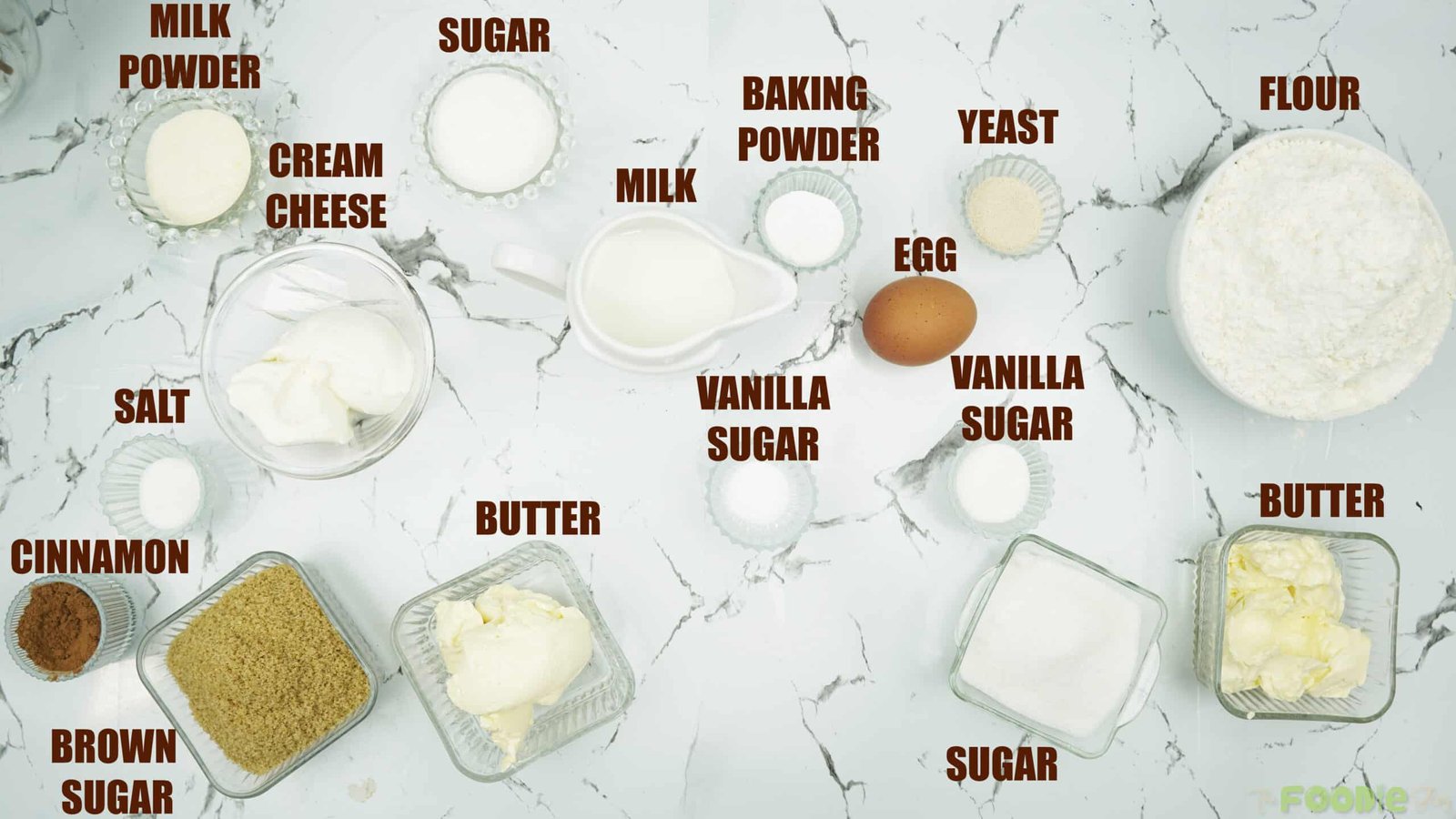 Flat lay of cinnamon roll ingredients including flour, yeast, milk, butter, egg, sugar, cinnamon, cream cheese, baking powder, and milk powder