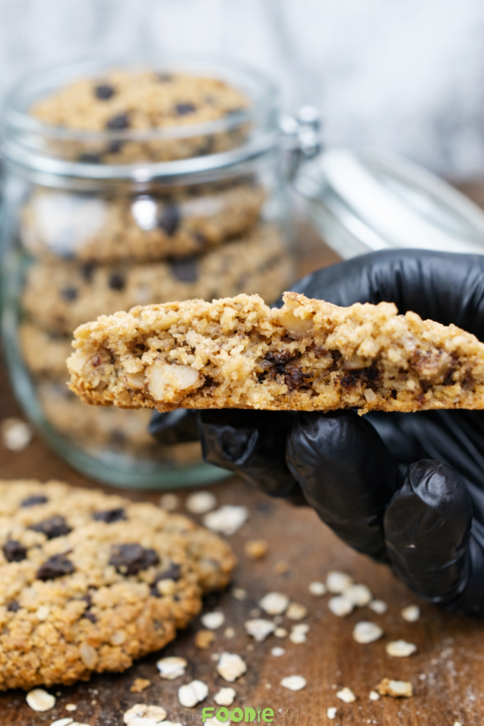 Half of a classic walnut cookie held by hand, showing the inside texture with oats, chopped walnuts, and chocolate.