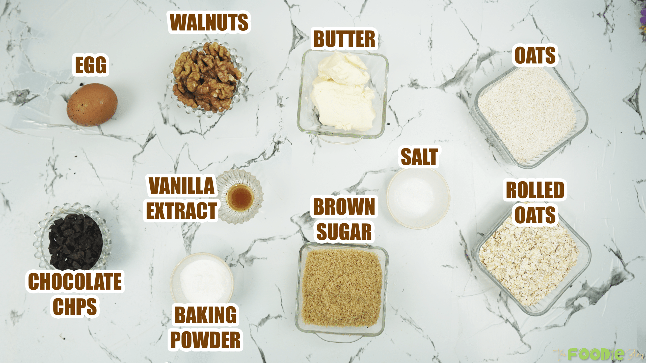 Labeled ingredients on a counter including oats, rolled oats, walnuts, butter, brown sugar, egg, baking powder, salt, and chocolate chips.