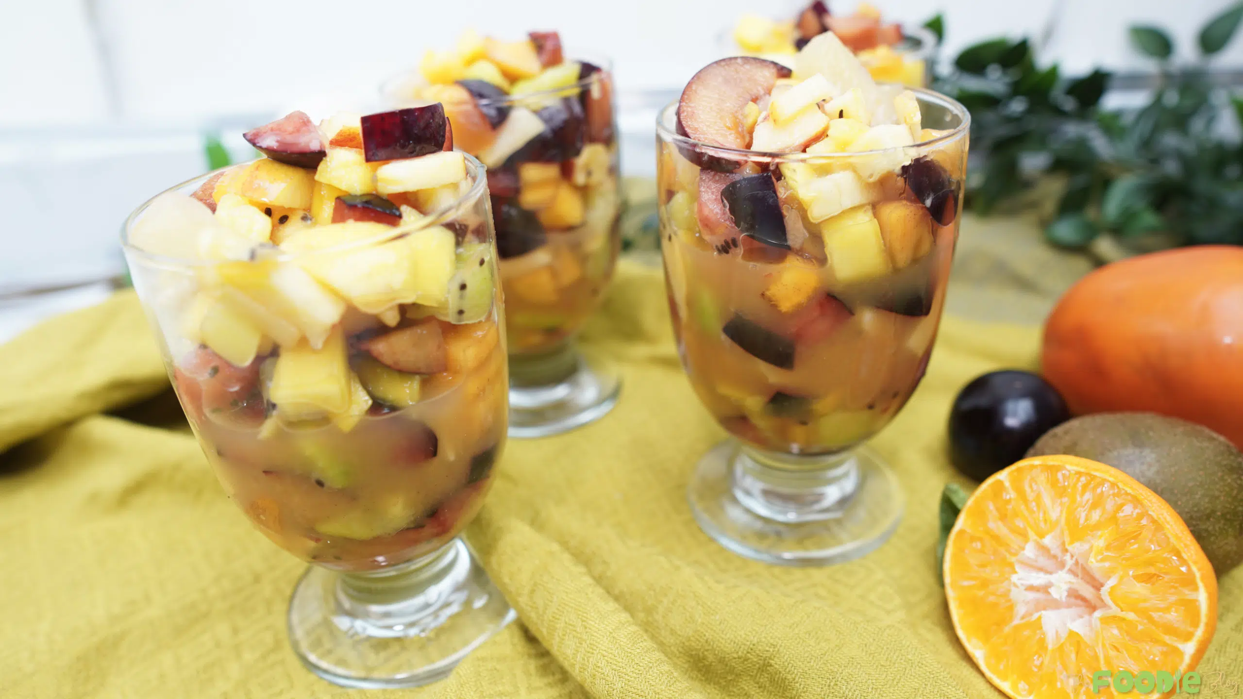 Three glasses of fruit salad with pineapple, kiwi, plums, and apples on a yellow cloth