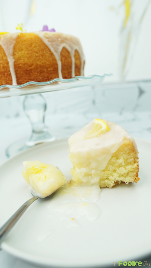 Slice of lemon cake topped with lemon glaze on a white plate, with the glazed cake on a stand in the background