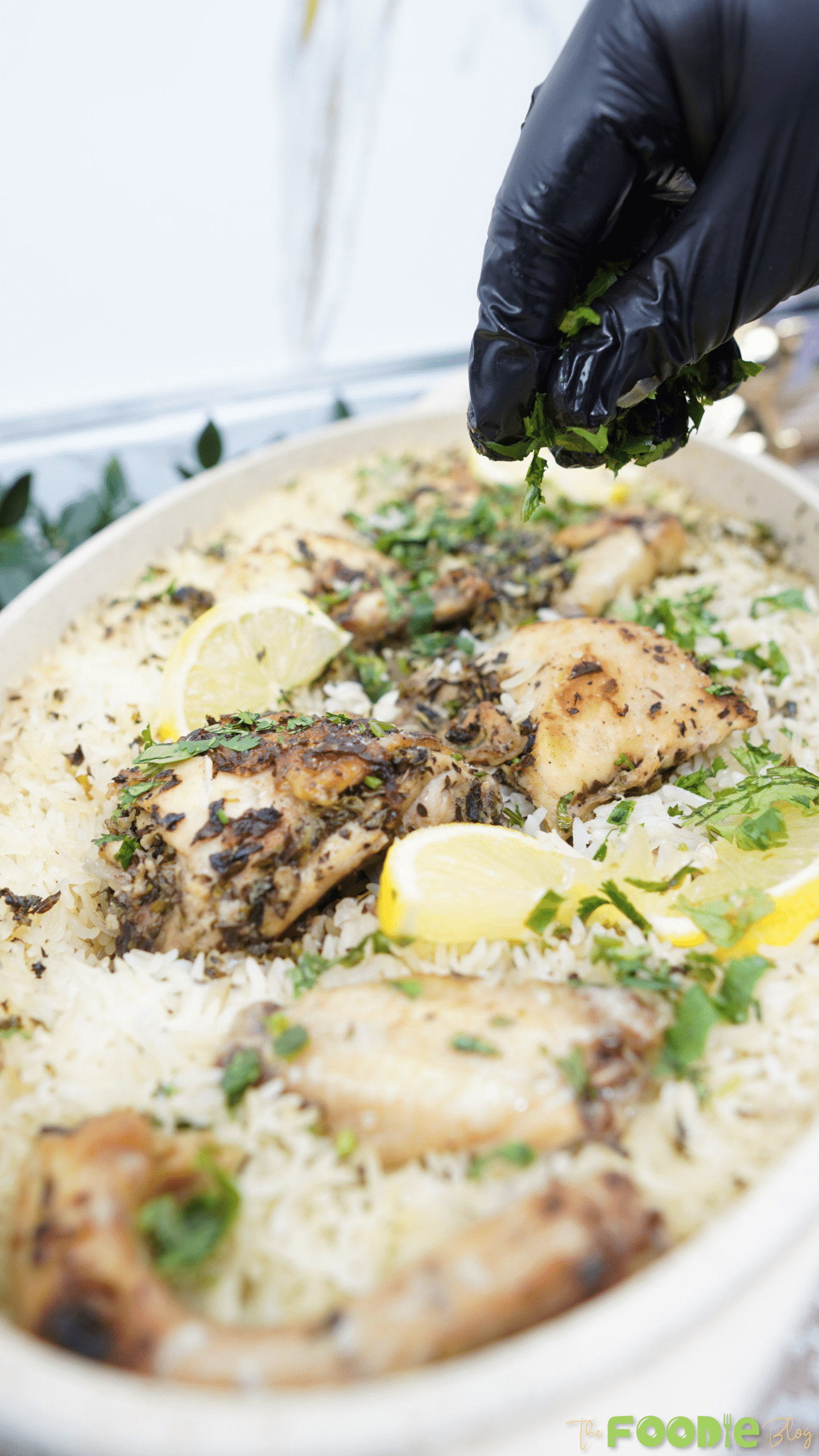 Gloved hand sprinkling chopped parsley over one pot chicken and lemon rice with lemon slices
