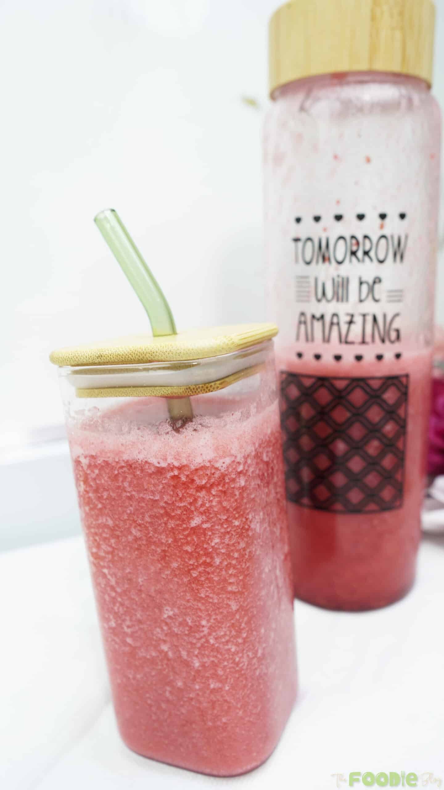 Iced raspberry juice in a square glass with a straw, with a bottle of juice in the background