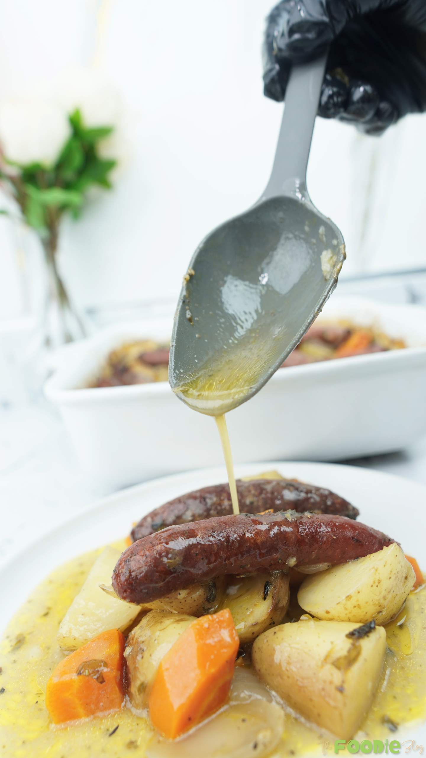 Spoon drizzling gravy over a plated sausage bake with potatoes, carrots, and onions