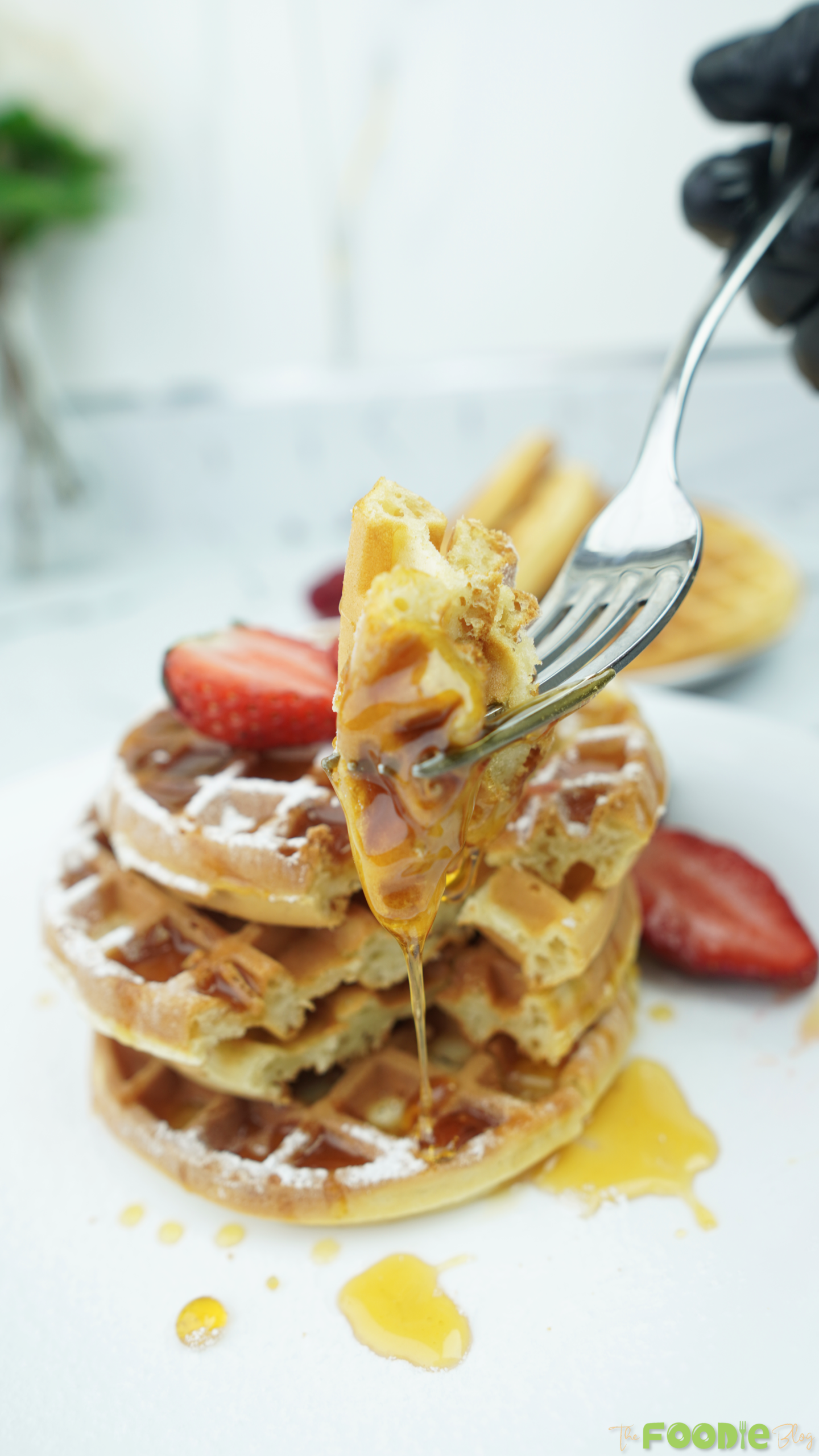 Fork holding a bite of mini waffle with syrup dripping in front of a waffle stack