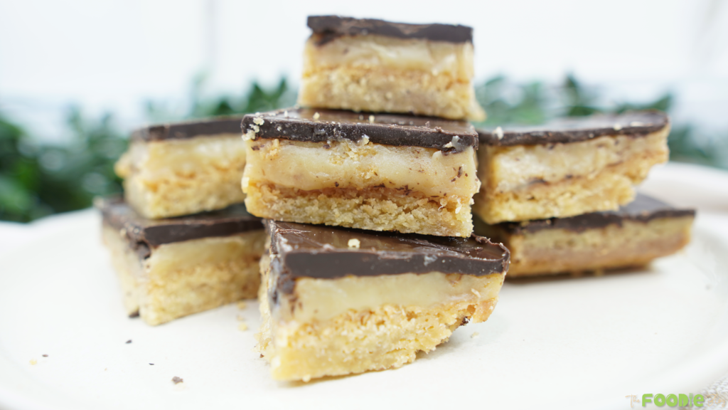 Stacked caramel slice squares with a chocolate top layer and caramel filling on a white plate