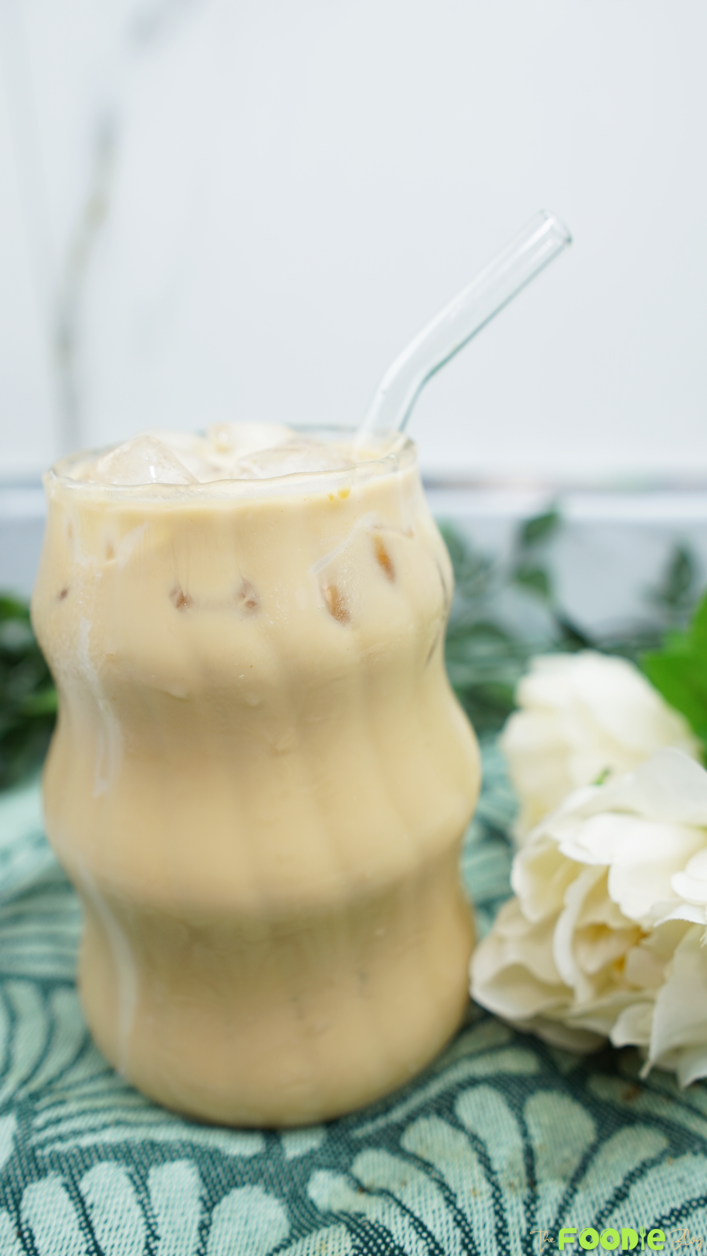 Iced coffee with milk in a glass, served with a straw and flowers in the background
