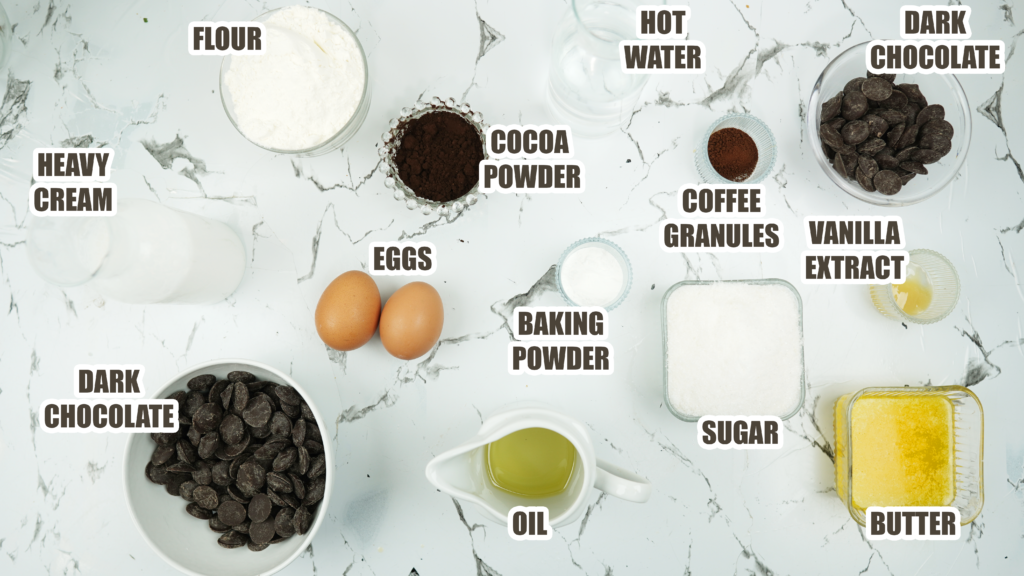 Overhead view of ingredients for Chocolate Fudge Cake, including flour, cocoa powder, dark chocolate, eggs, sugar, butter, oil, hot water, coffee granules, vanilla, baking powder, and heavy cream.