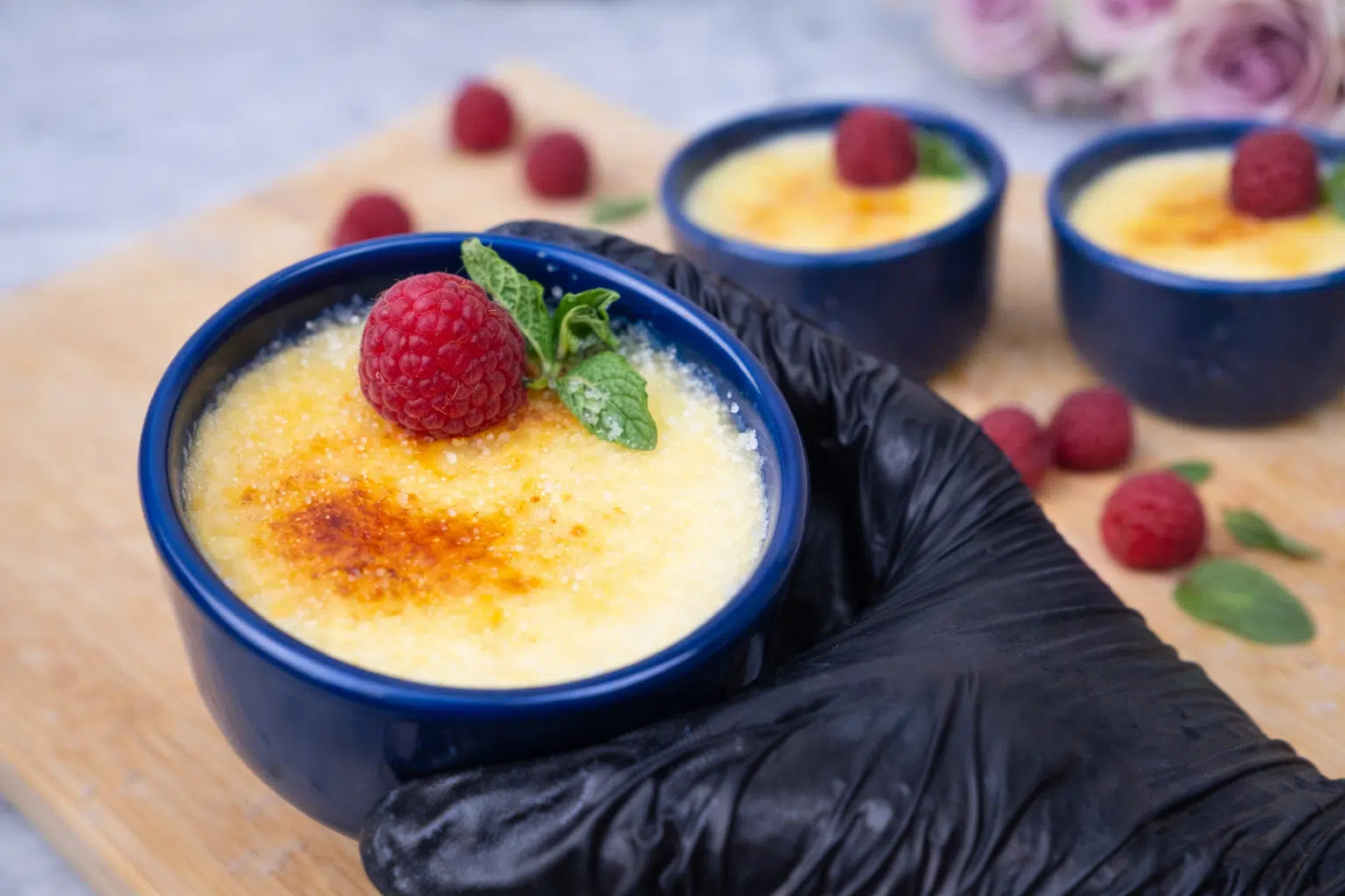 Close-up of creme brulee in a blue ramekin topped with raspberries and mint on a wooden board.