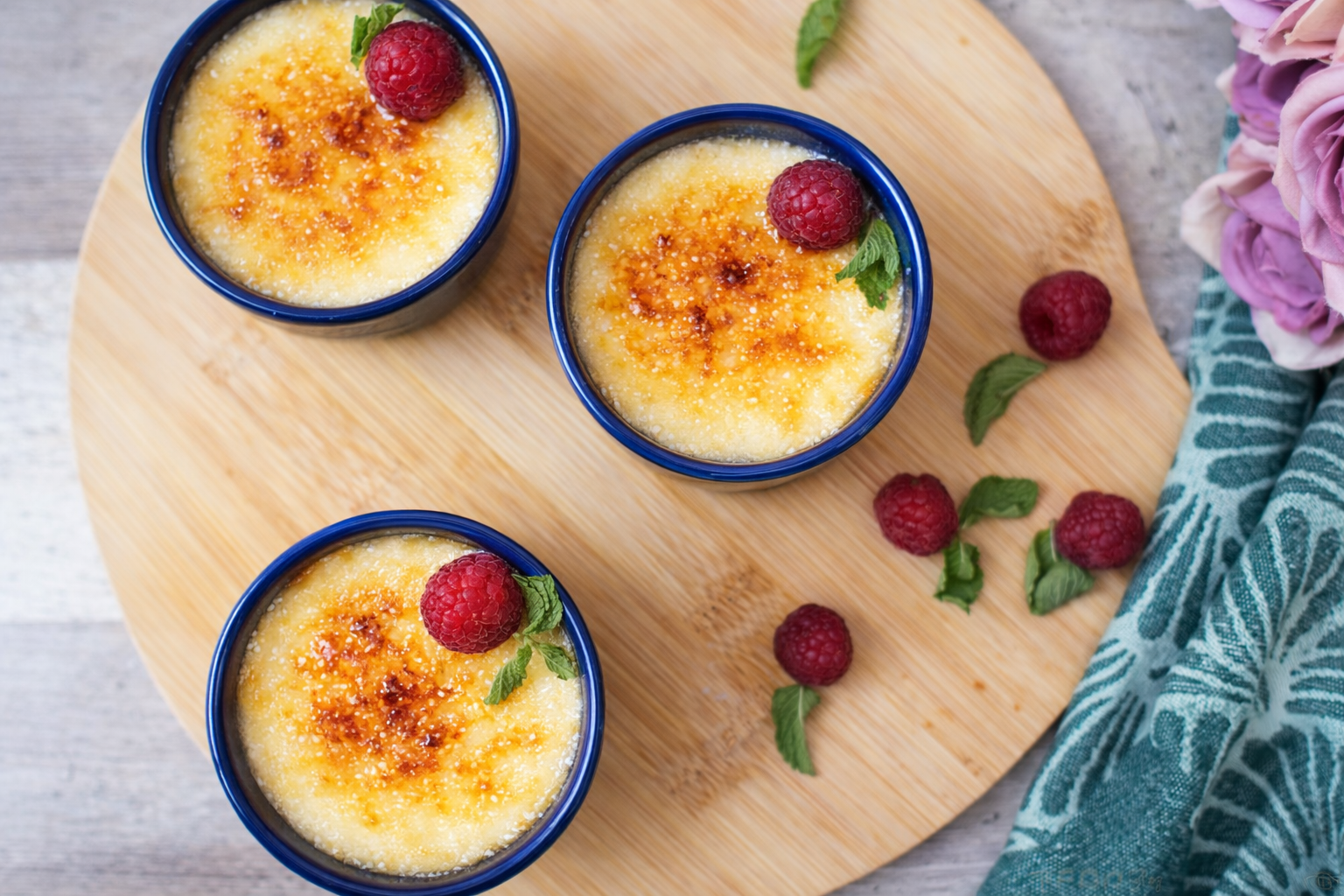 Overhead view of three creme brulee ramekins topped with raspberries and mint on a round wooden board.