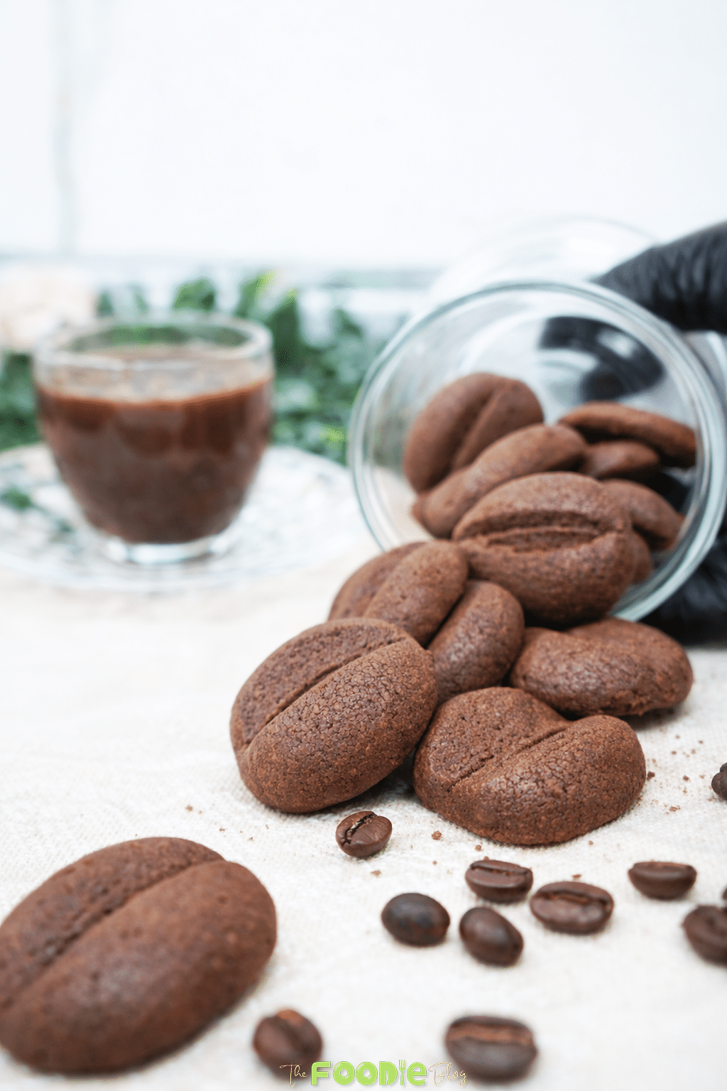 Crispy coffee biscuits shaped like coffee beans served beside a glass of coffee with scattered roasted coffee beans.