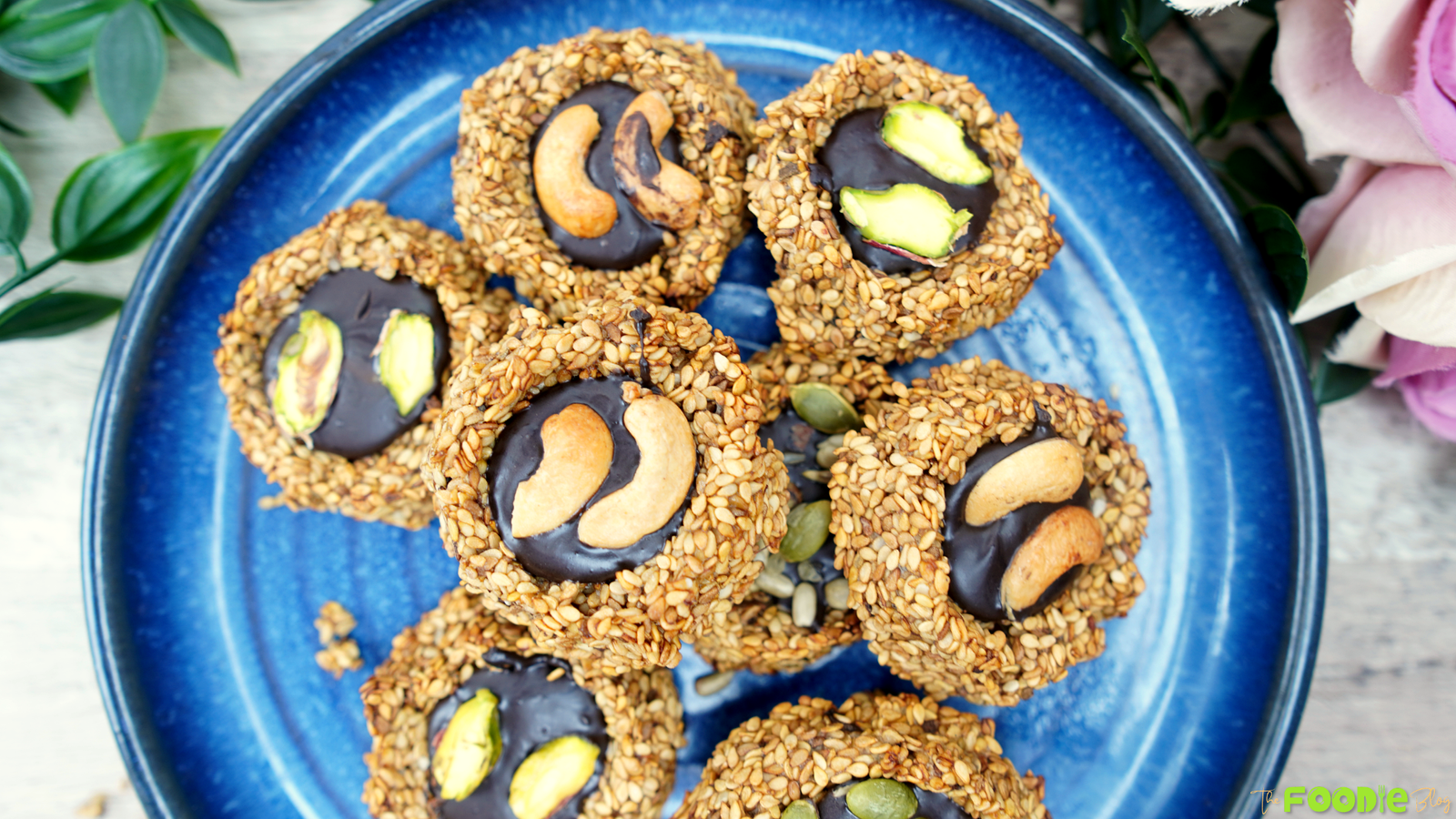 Overhead view of sesame-coated banana cookies with dark chocolate centers topped with cashews and pistachios on a blue plate