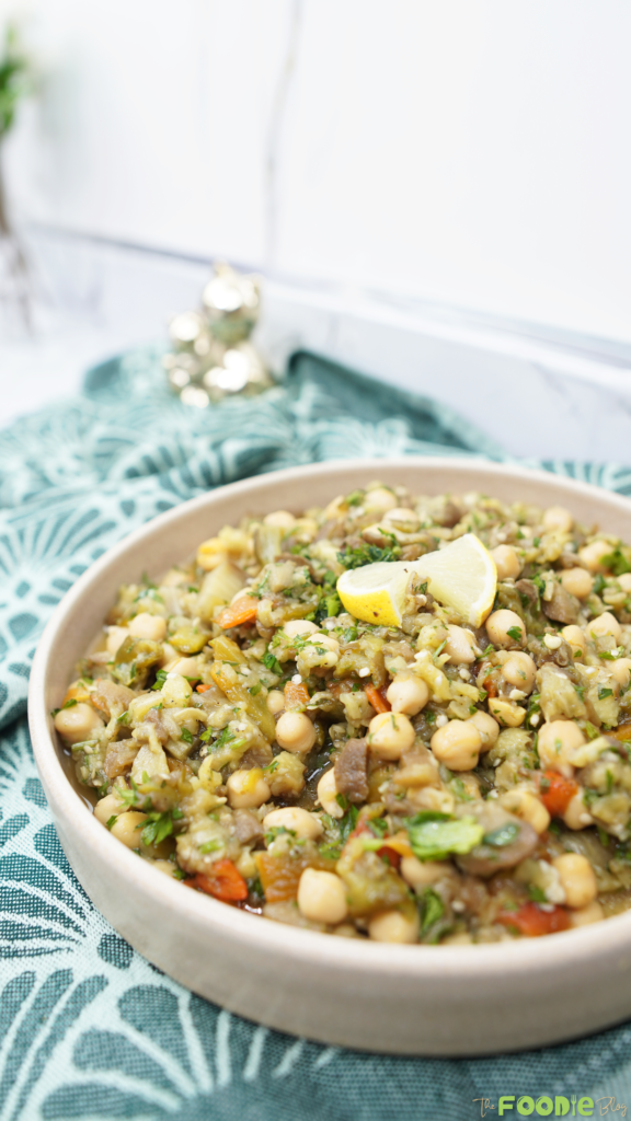 Bowl of eggplant chickpea salad on a teal cloth with lemon wedges and a bright background