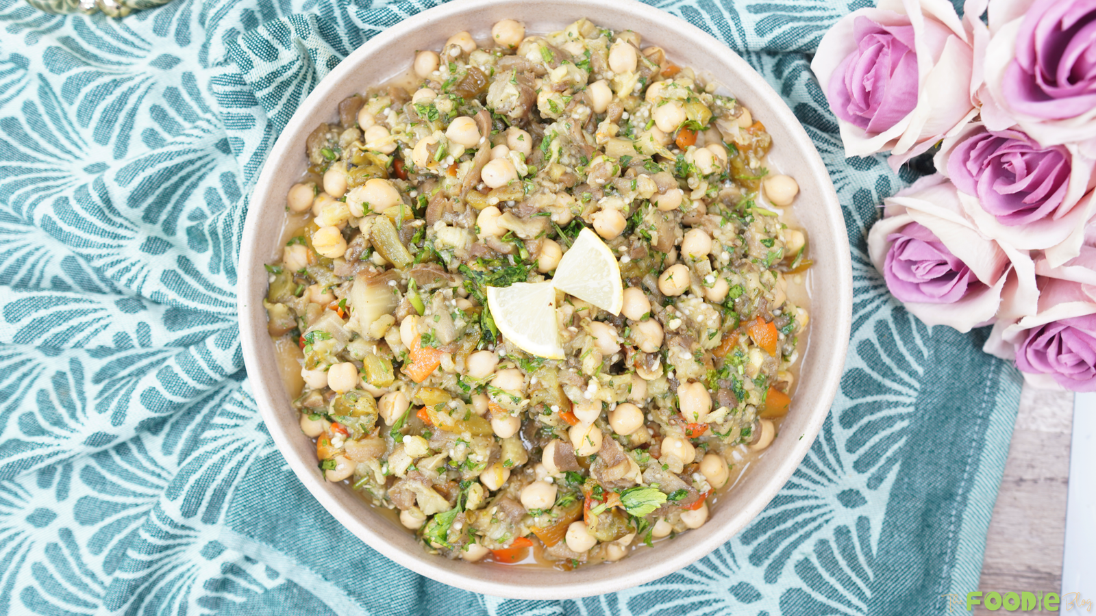 Overhead view of eggplant chickpea salad with lemon wedges in a beige bowl on a teal cloth