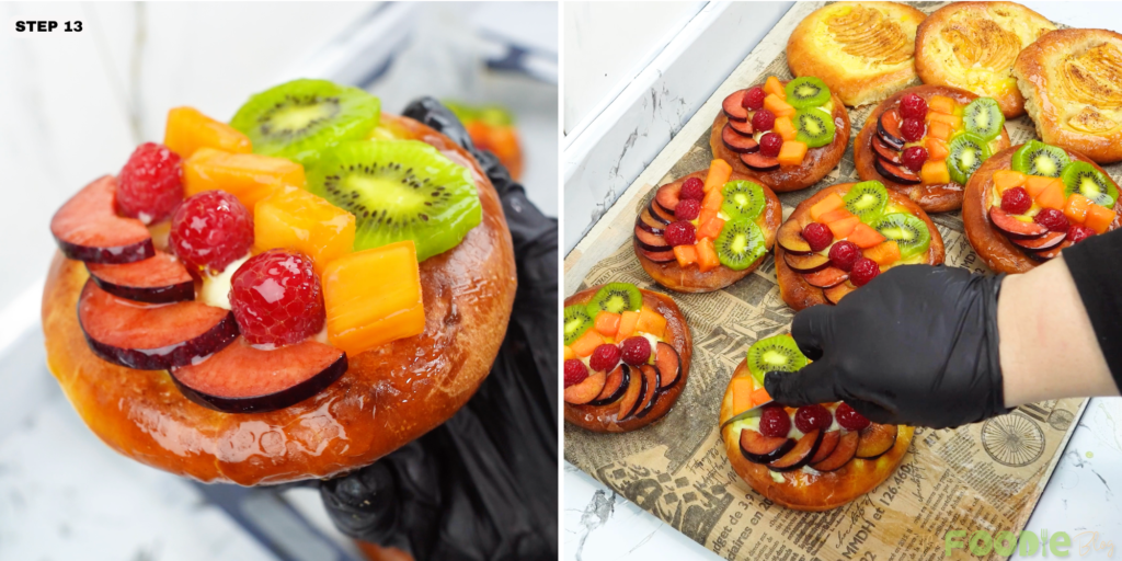 A hand places kiwi and other fruit on pastry-cream-topped brioche pastries on a tray.