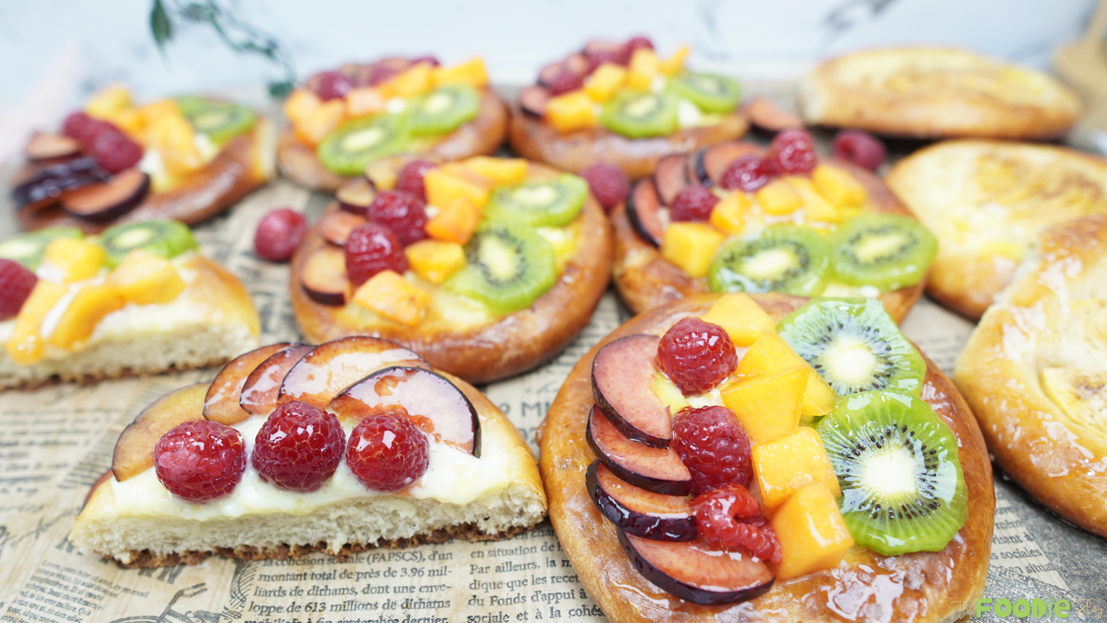 A tray of glossy brioche topped with pastry cream and fresh fruit like kiwi, plums, persimmon, and berries.