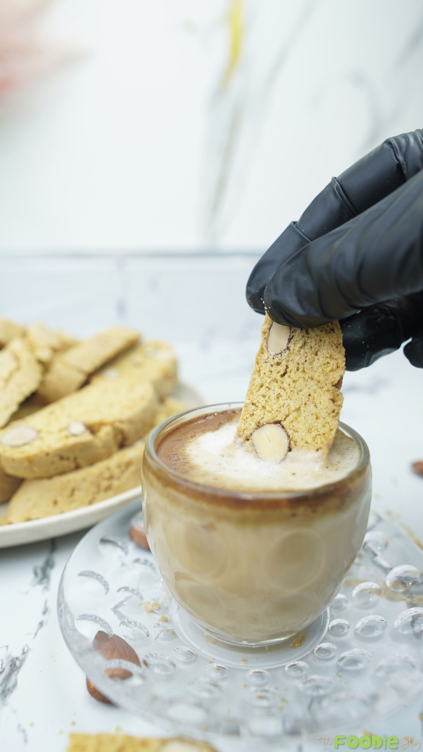 A hand dipping a homemade almond biscotti into a cup of coffee with biscotti on a plate in the background.