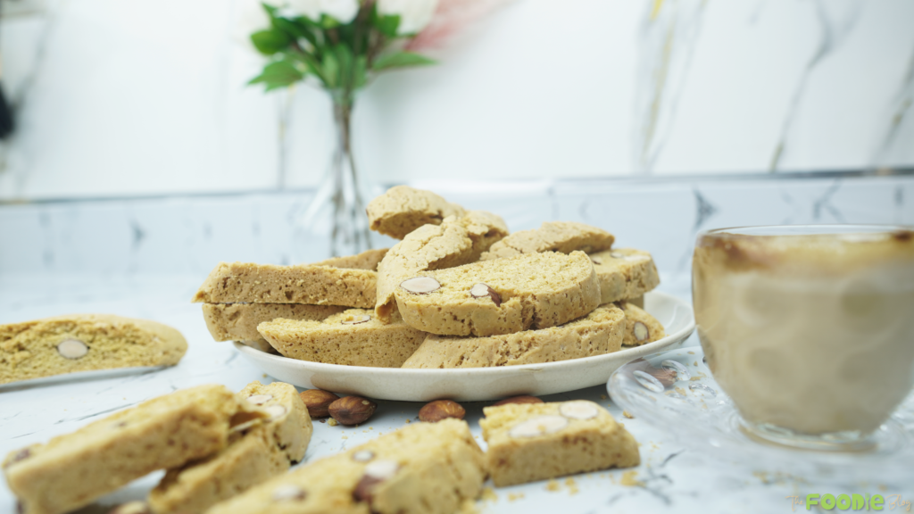 Homemade almond biscotti stacked on a plate with a coffee cup nearby and crumbs on the counter.