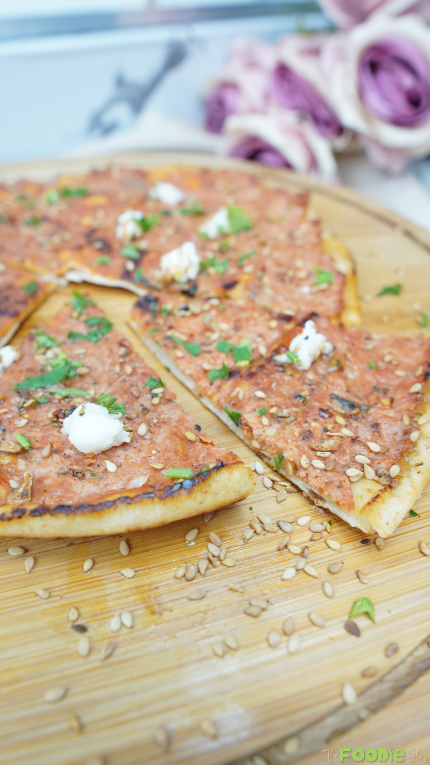 A hand holding a slice of Lebanese flatbread pizza topped with sesame seeds, parsley, and goat cheese