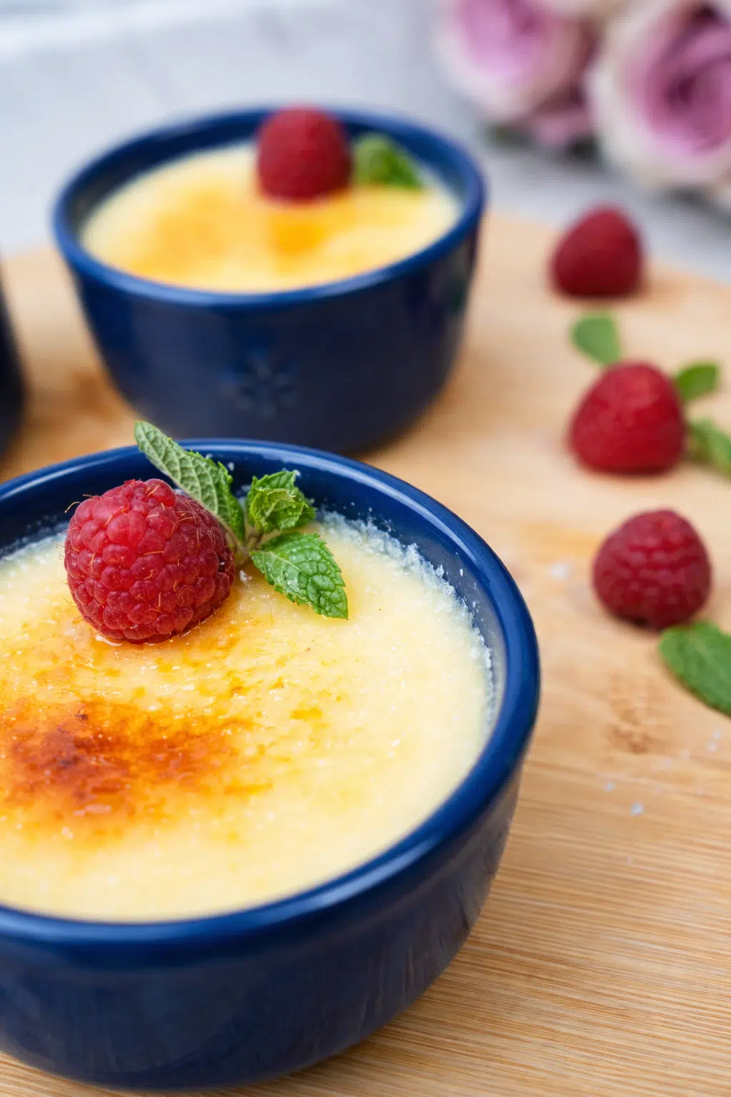 Close-up of creme brulee in a blue ramekin topped with raspberries and mint on a wooden board.