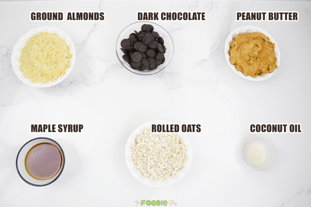 Ground almonds, dark chocolate, peanut butter, maple syrup, rolled oats, and coconut oil arranged on a light surface
