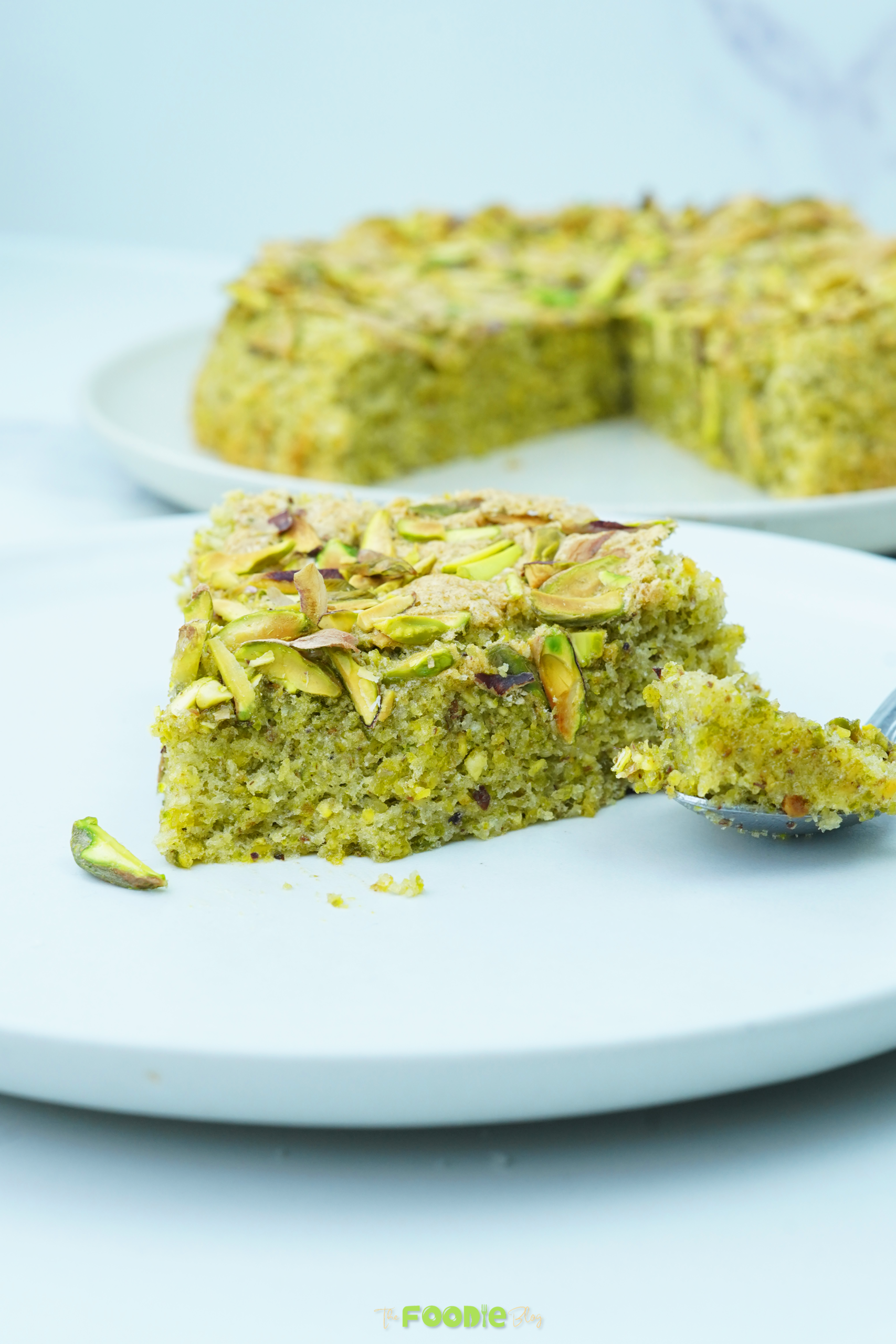 Slice of pistachio cake on a white plate with the rest of the cake in the background