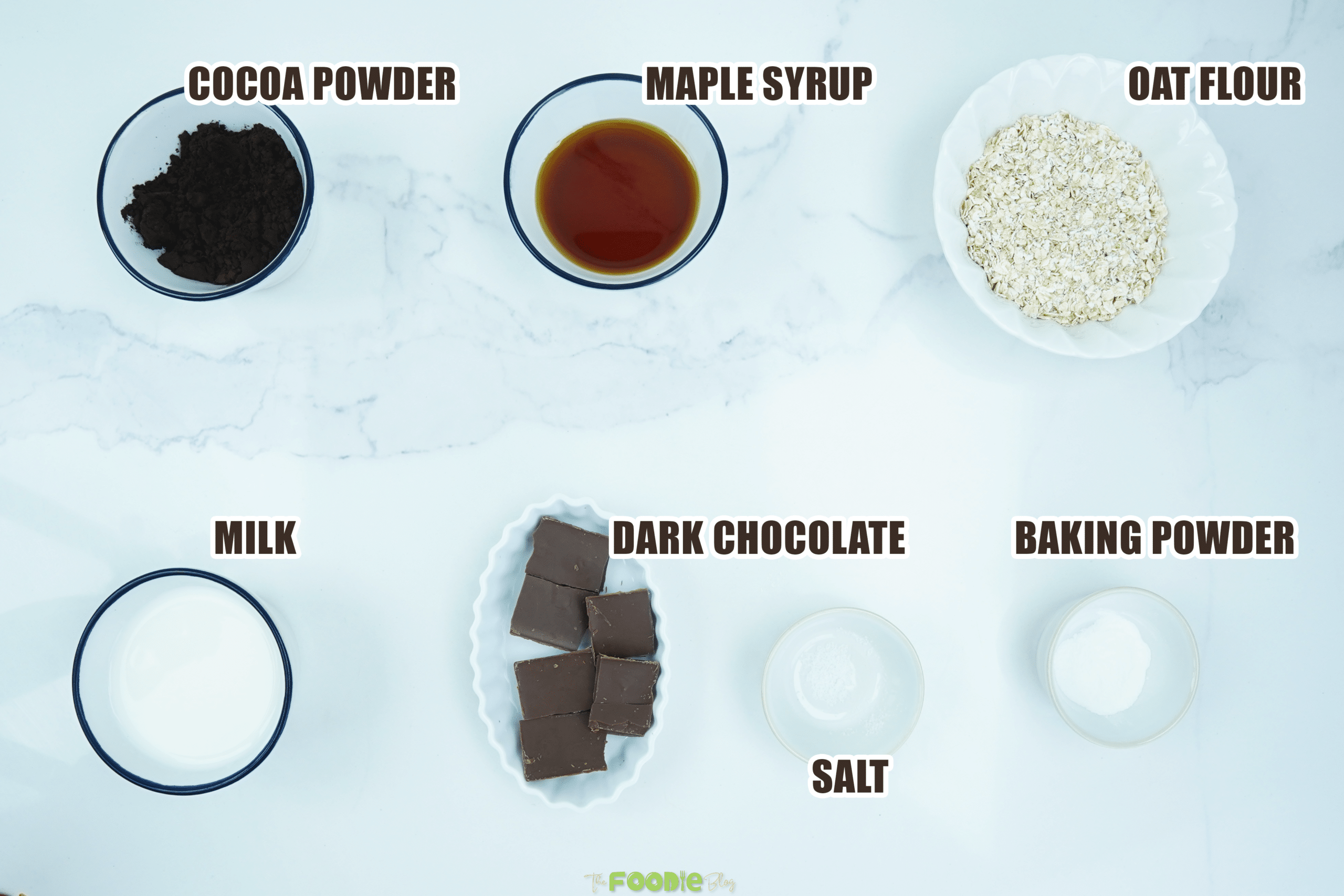 Oat flour, cocoa powder, maple syrup, milk, dark chocolate, baking powder, and salt on a white surface