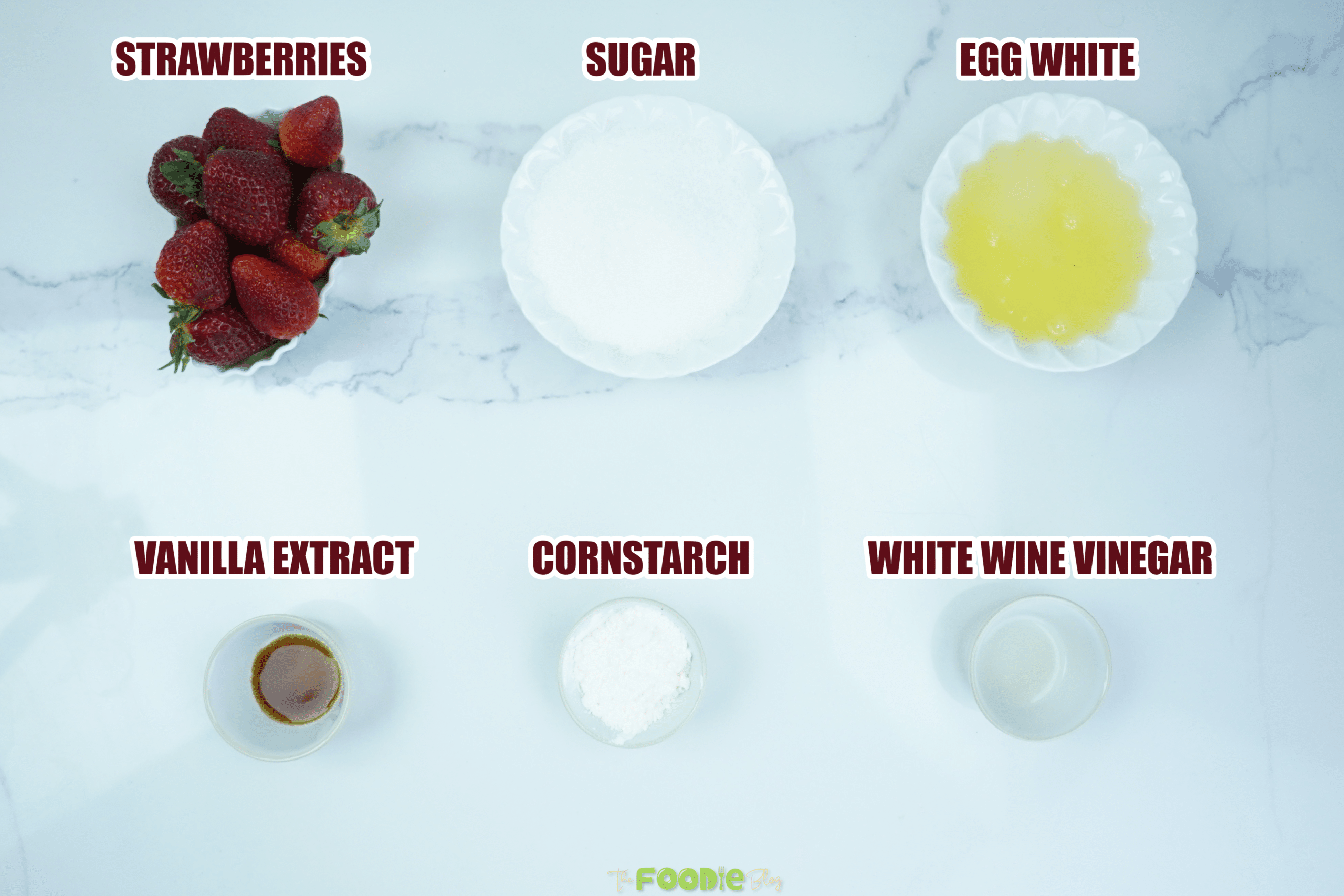 Ingredients for pavlova arranged on a light surface, including strawberries, sugar, egg whites, cornstarch, vinegar, and vanilla