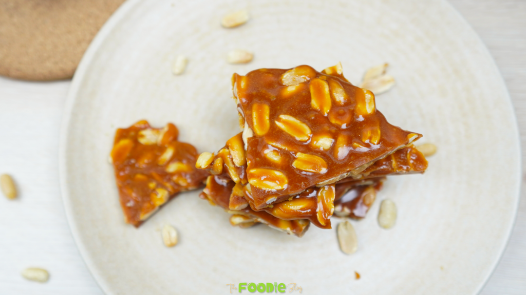 Peanut brittle pieces stacked on a plate with a few peanuts scattered around Caption: Finished peanut brittle ready to serve