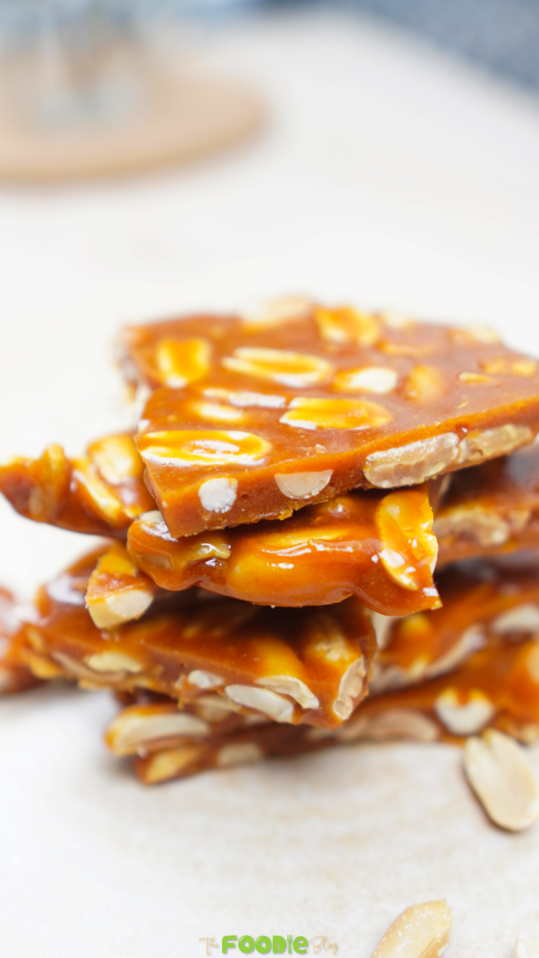 Stack of glossy peanut brittle pieces with peanuts around it Caption: Crisp, buttery peanut brittle with roasted peanuts
