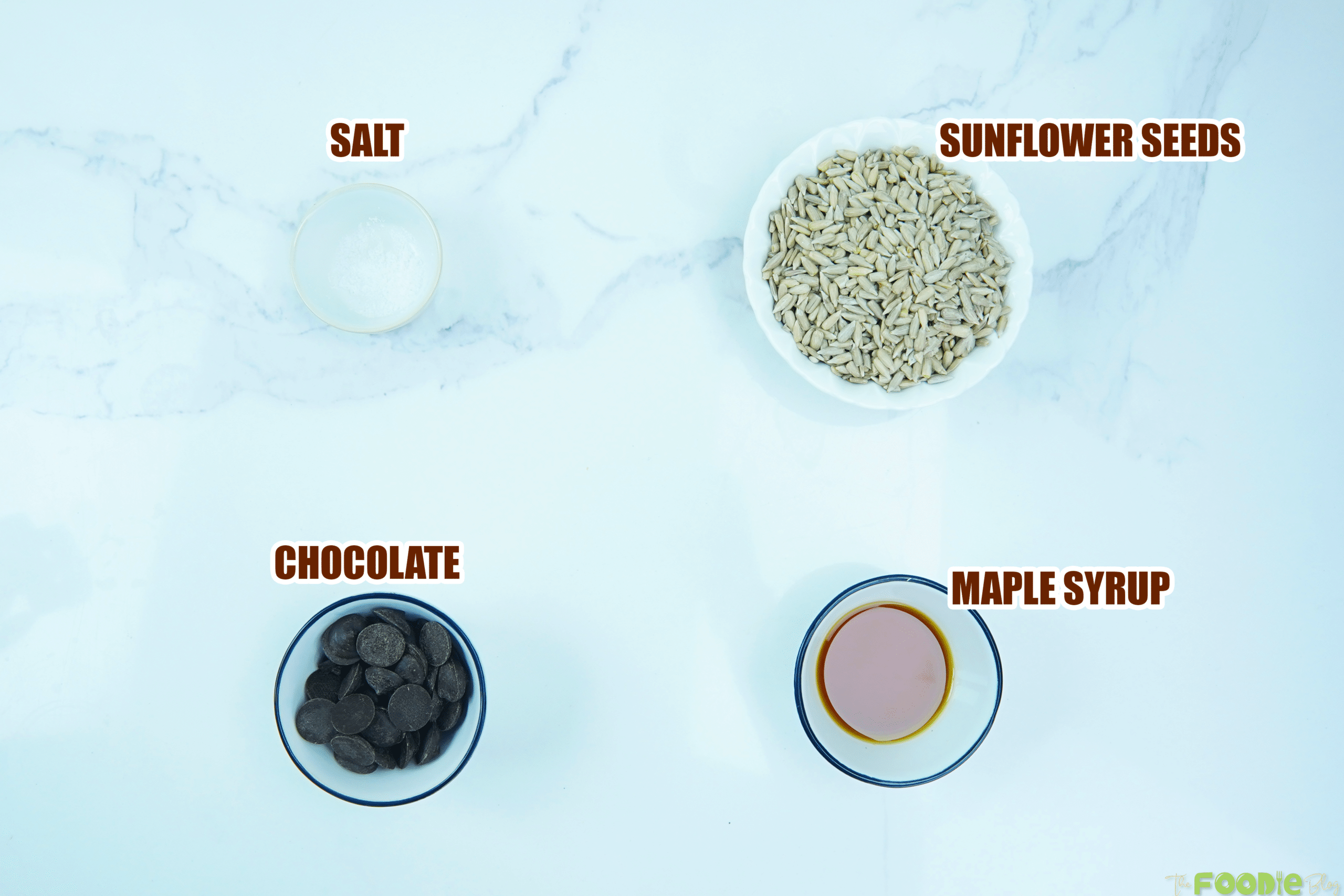Bowls of sunflower seeds, chocolate, maple syrup, and salt on a light surface