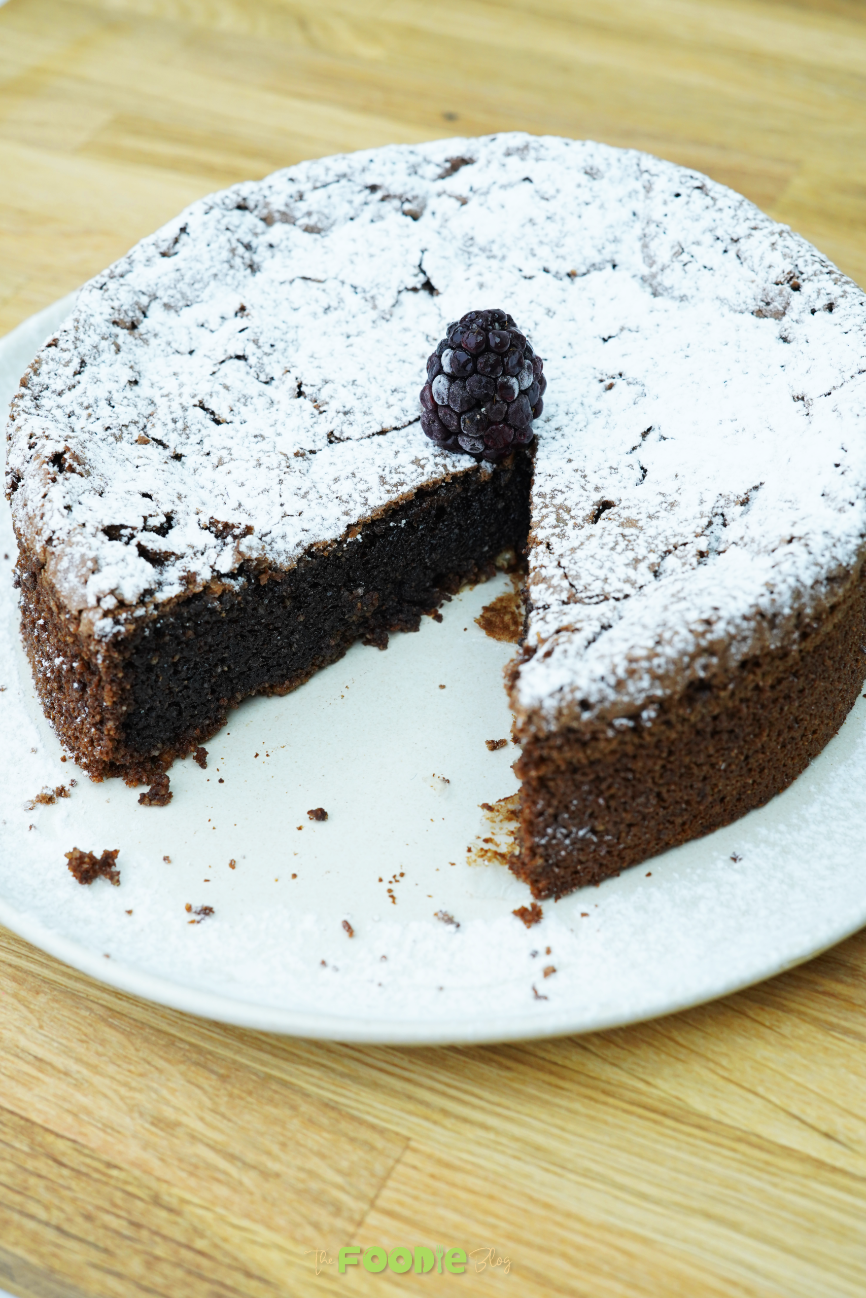 Whole torta caprese cake dusted with powdered sugar with one slice removed and a blackberry on top