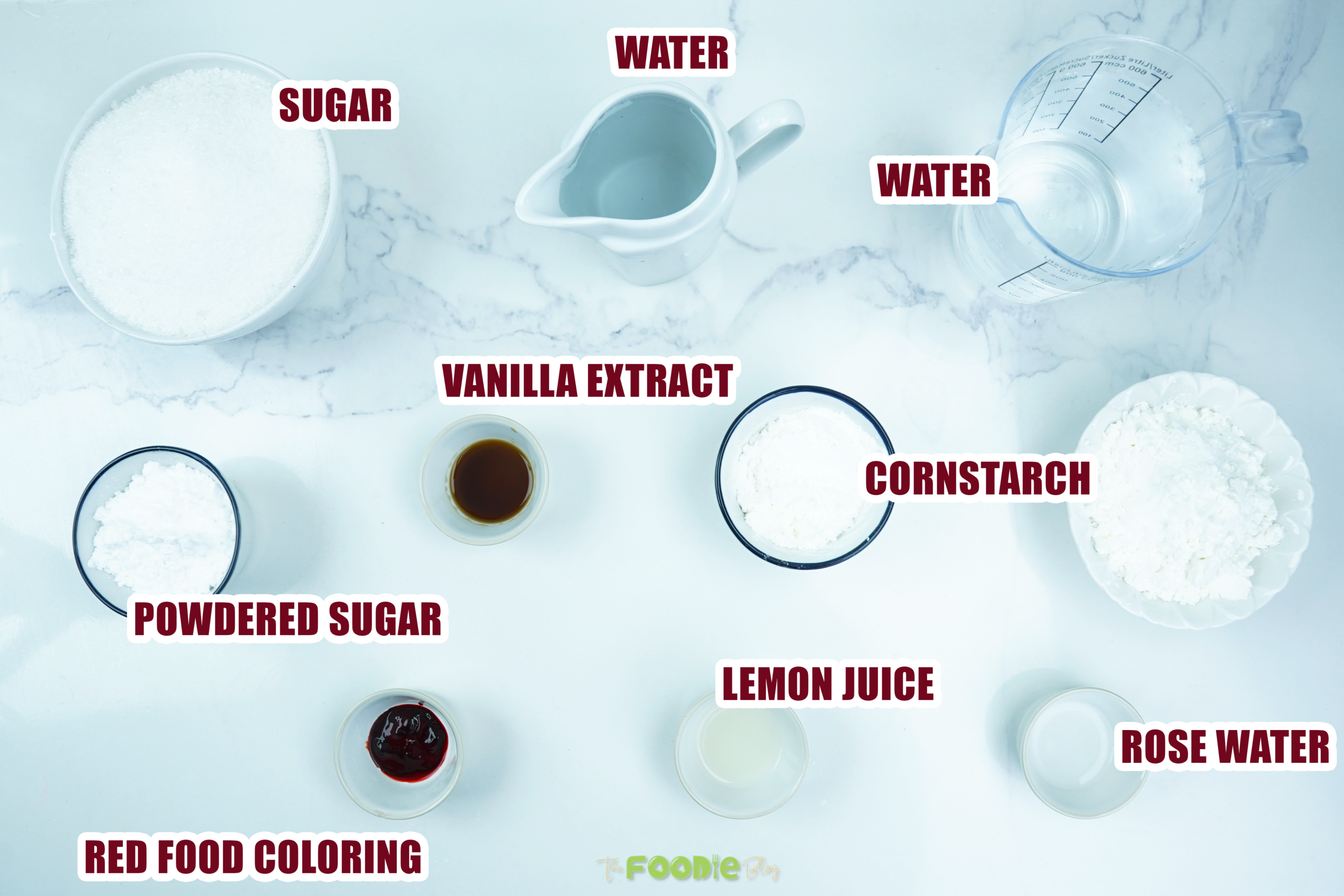 Overhead view of sugar, water, vanilla, lemon juice, rose water, cornstarch, powdered sugar, and red coloring labeled on a counter