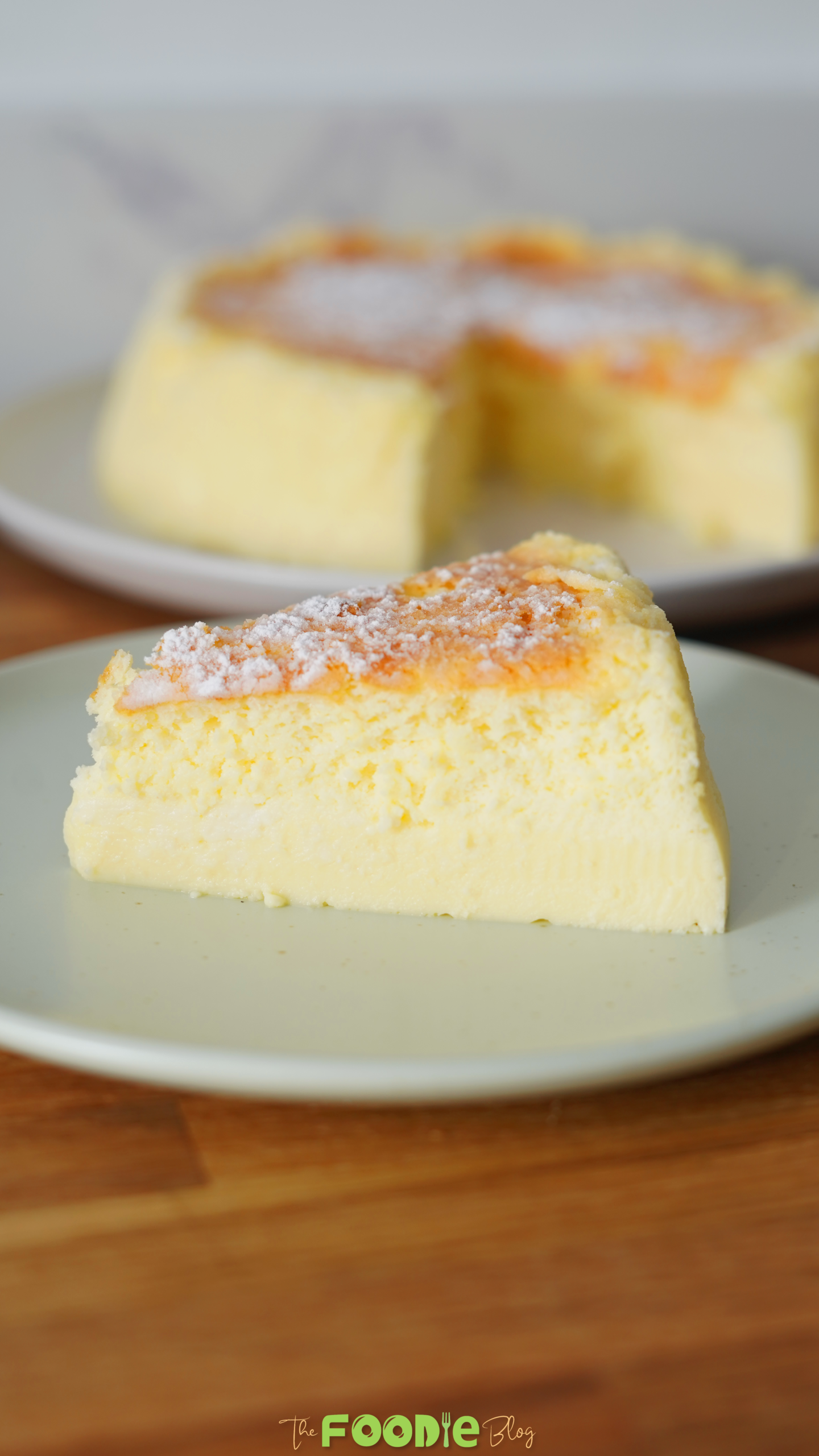 Single slice of Japanese cheesecake dusted with powdered sugar on a plate with the rest of the cake blurred in the background