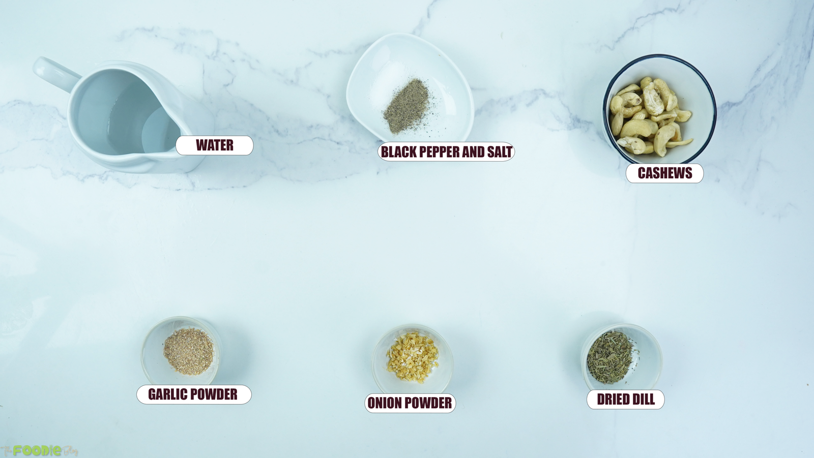 Ingredients for cashew ranch dressing arranged on a white marble surface, including cashews, water, dill, garlic powder, onion powder, salt, and black pepper.