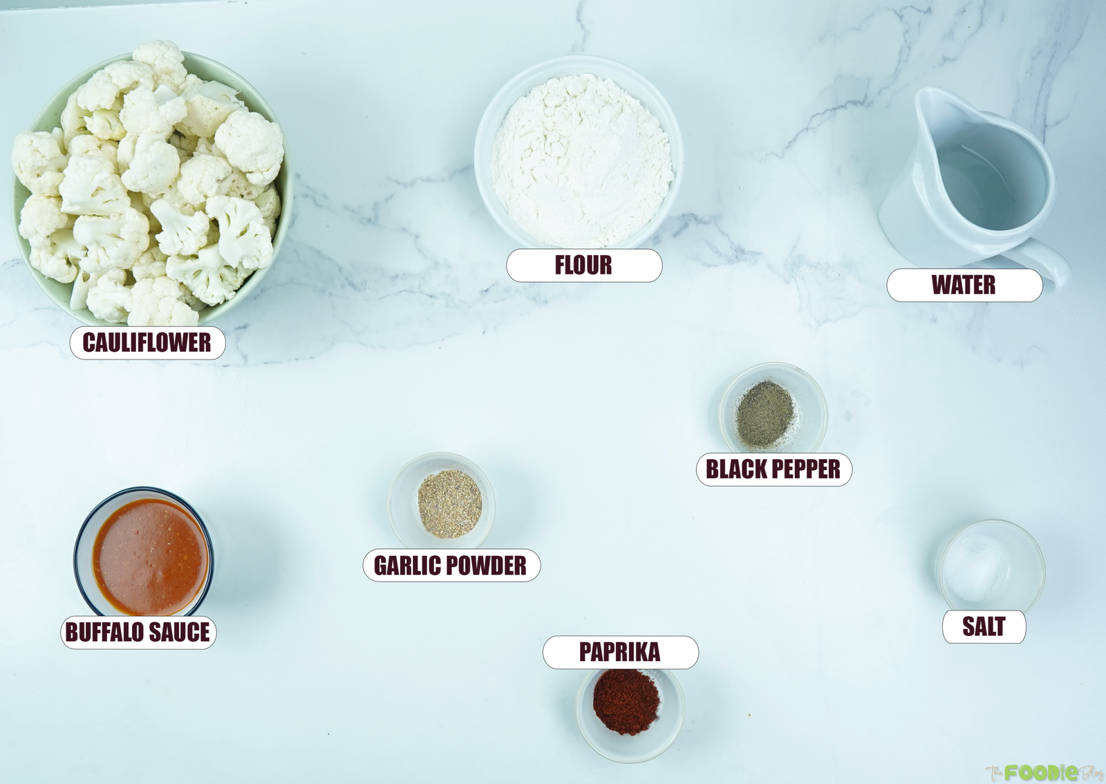 Ingredients for Buffalo Cauliflower Wings arranged on a white marble surface, including cauliflower, flour, water, buffalo sauce, garlic powder, paprika, salt, and black pepper.